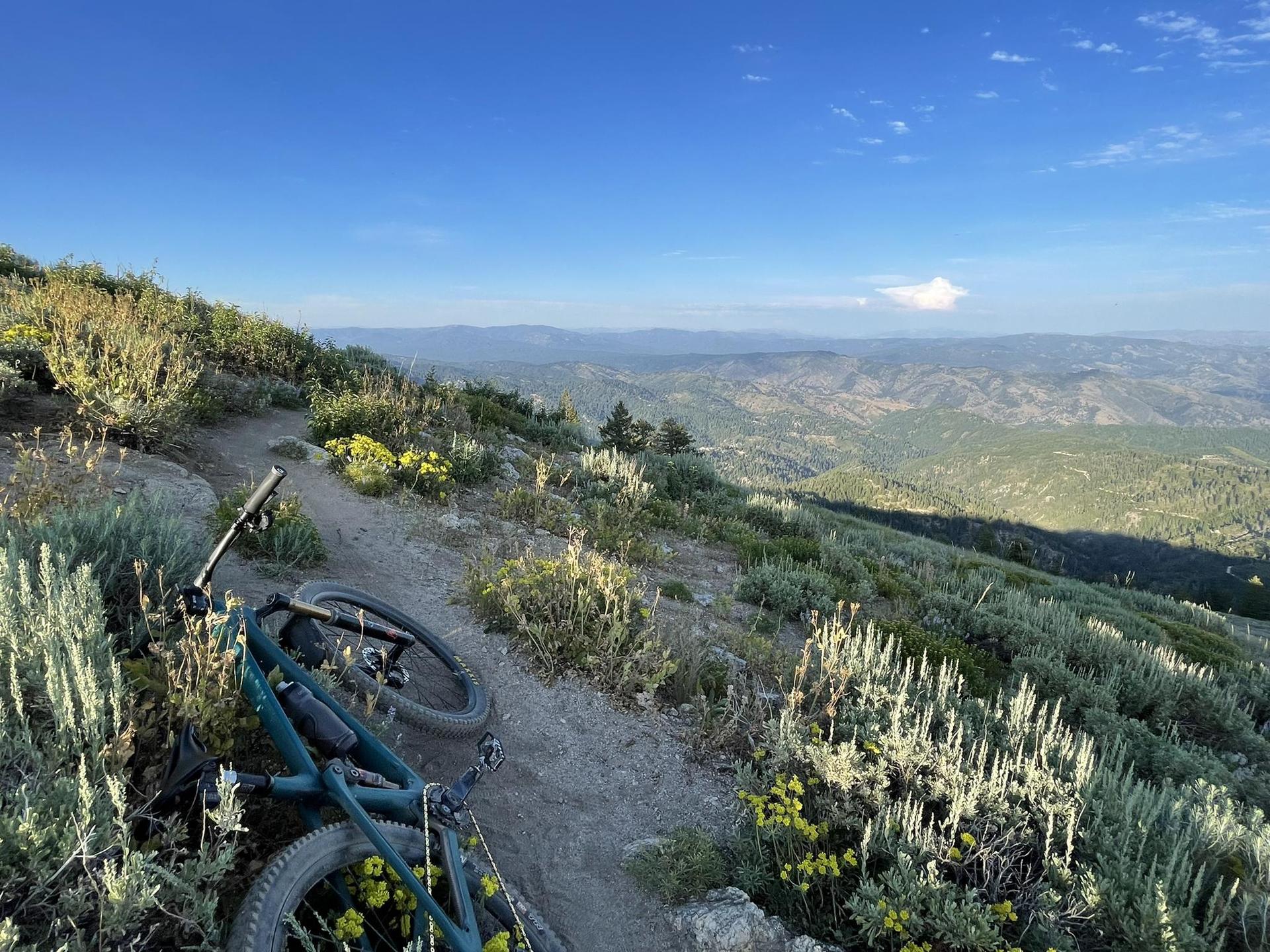 A mountain bike rests on a sunny trail with expansive mountain views.