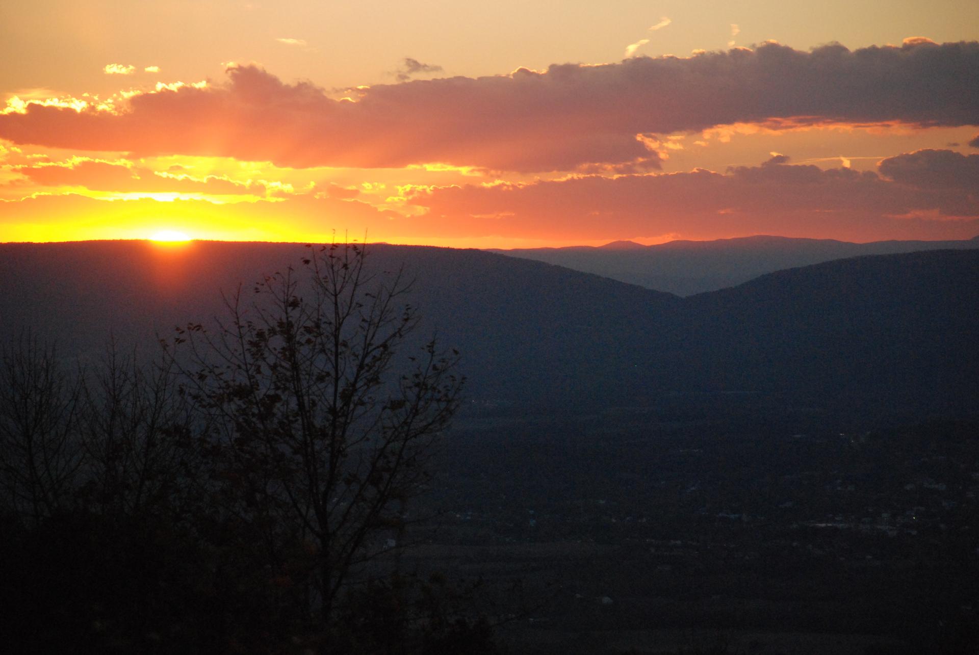 Golden sunset over dark mountain silhouettes with bare trees in the foreground.