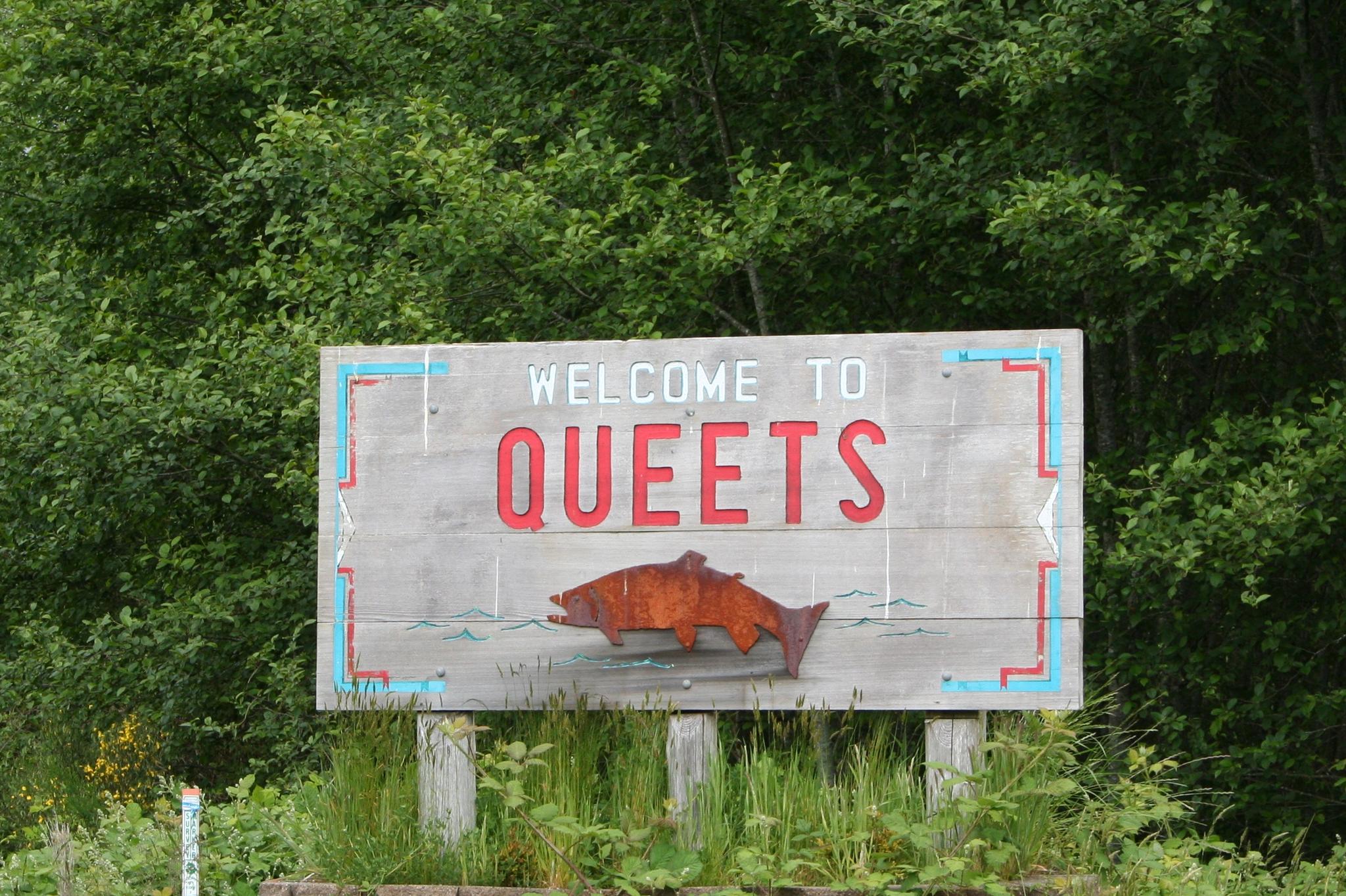 A rustic wooden welcome sign with a fish carving, surrounded by green trees.