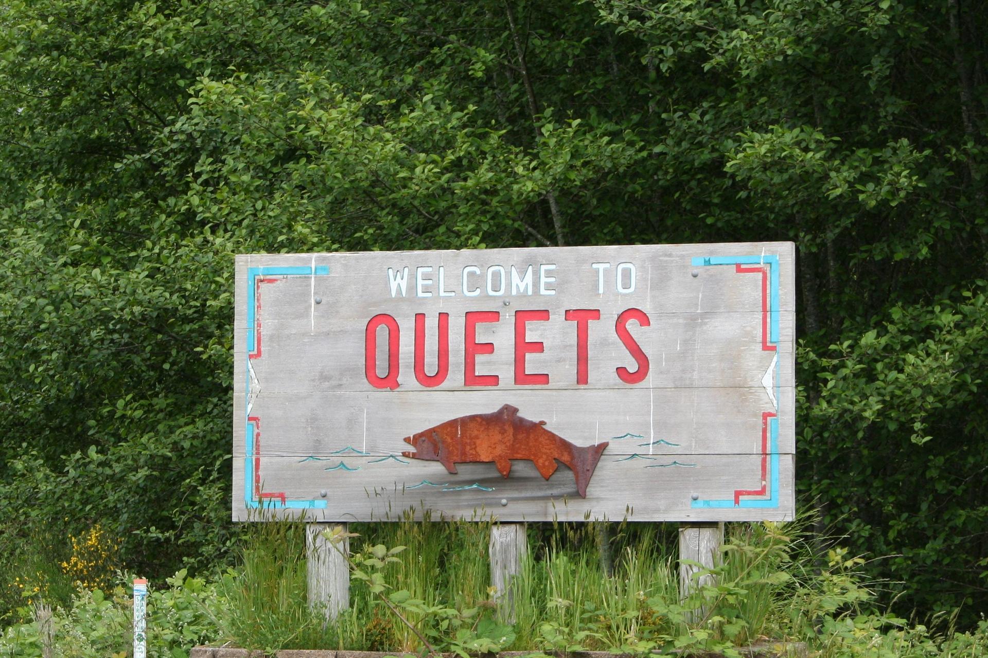 A rustic wooden welcome sign with a fish carving, surrounded by green trees.