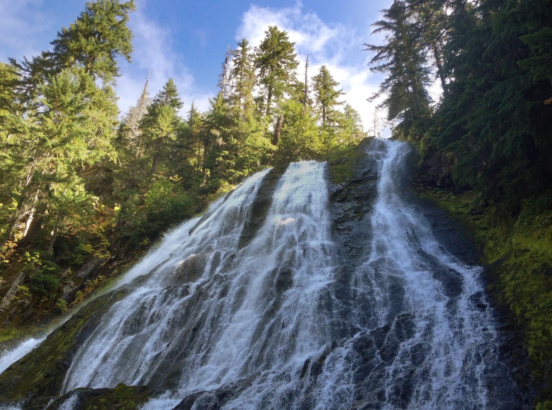 A powerful waterfall cascades down a mossy rock face under a blue sky.