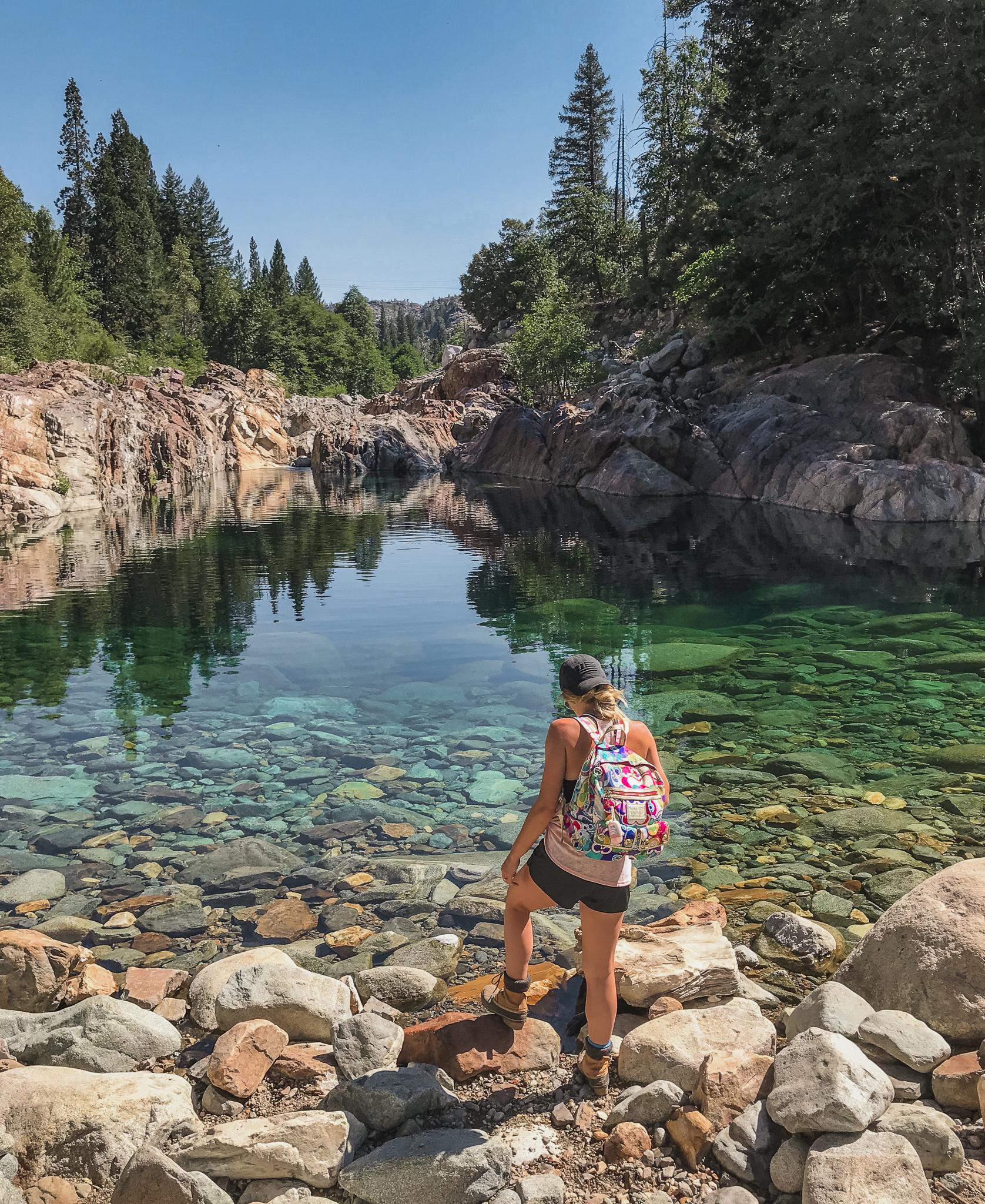 Person with backpack stands on rocks by clear green river, sunny day.