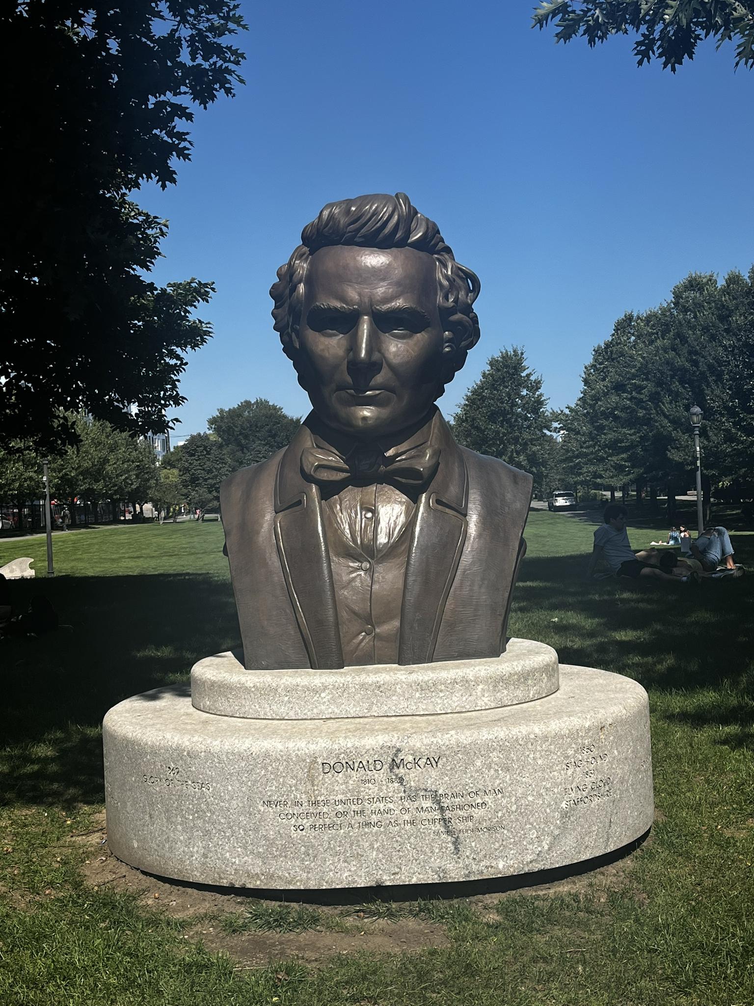 Bronze bust of a man on a pedestal in a sunny park with trees.