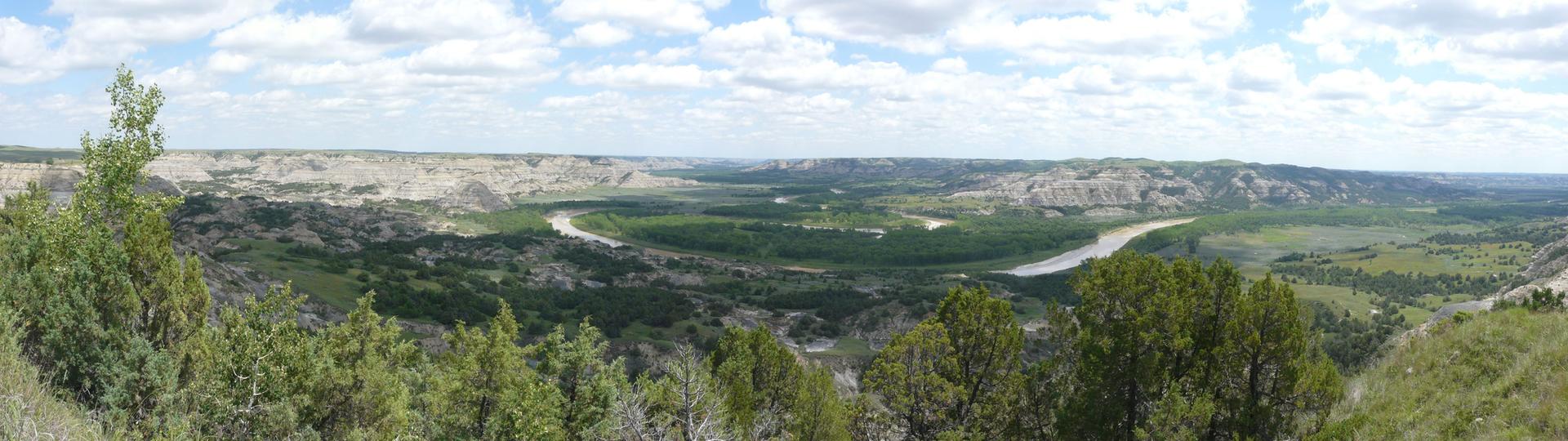 Panoramic view of a winding river through a green valley and badlands under a cloudy sky.
