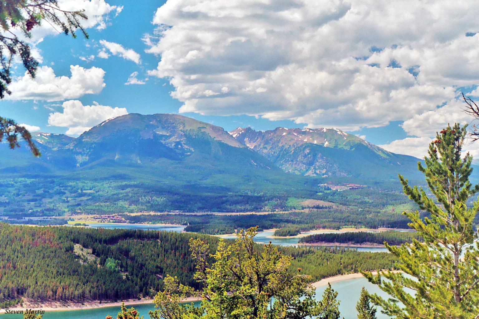 Panoramic view of a winding lake and forested mountains under a bright, cloudy sky.