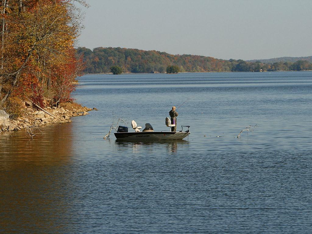 A person fishes from a small boat on a wide lake with autumn trees.