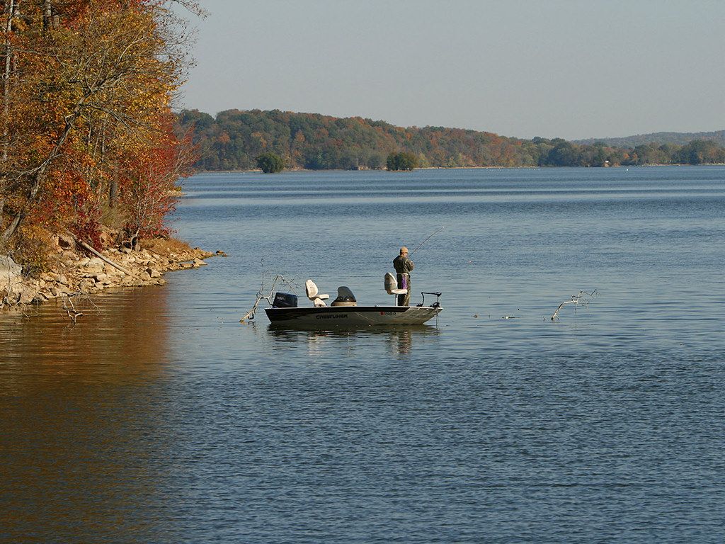 A person fishes from a small boat on a wide lake with autumn trees. - Central Hardwoods Scenic Trail