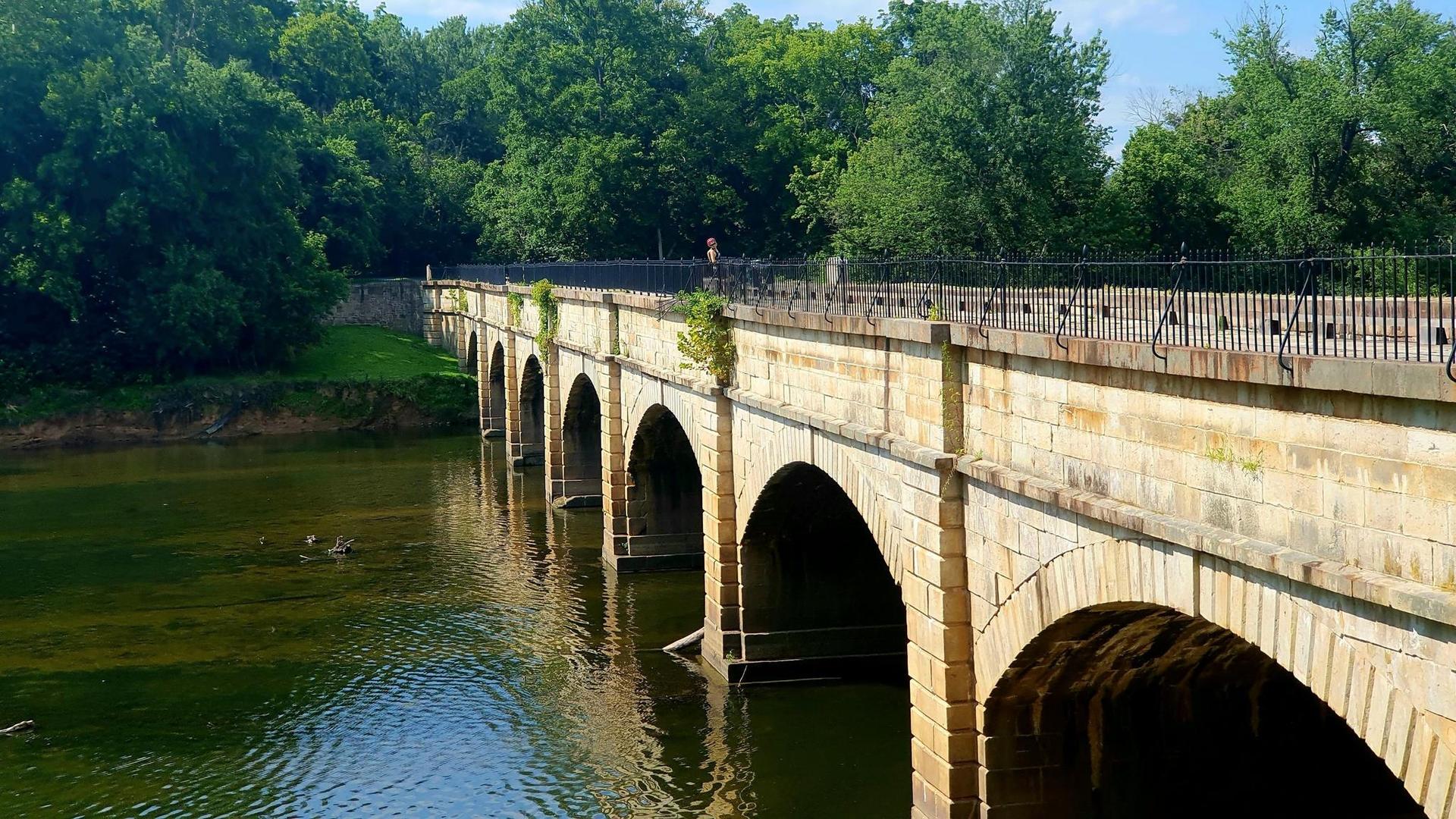 Stone arch bridge over water, surrounded by lush green trees on a sunny day.