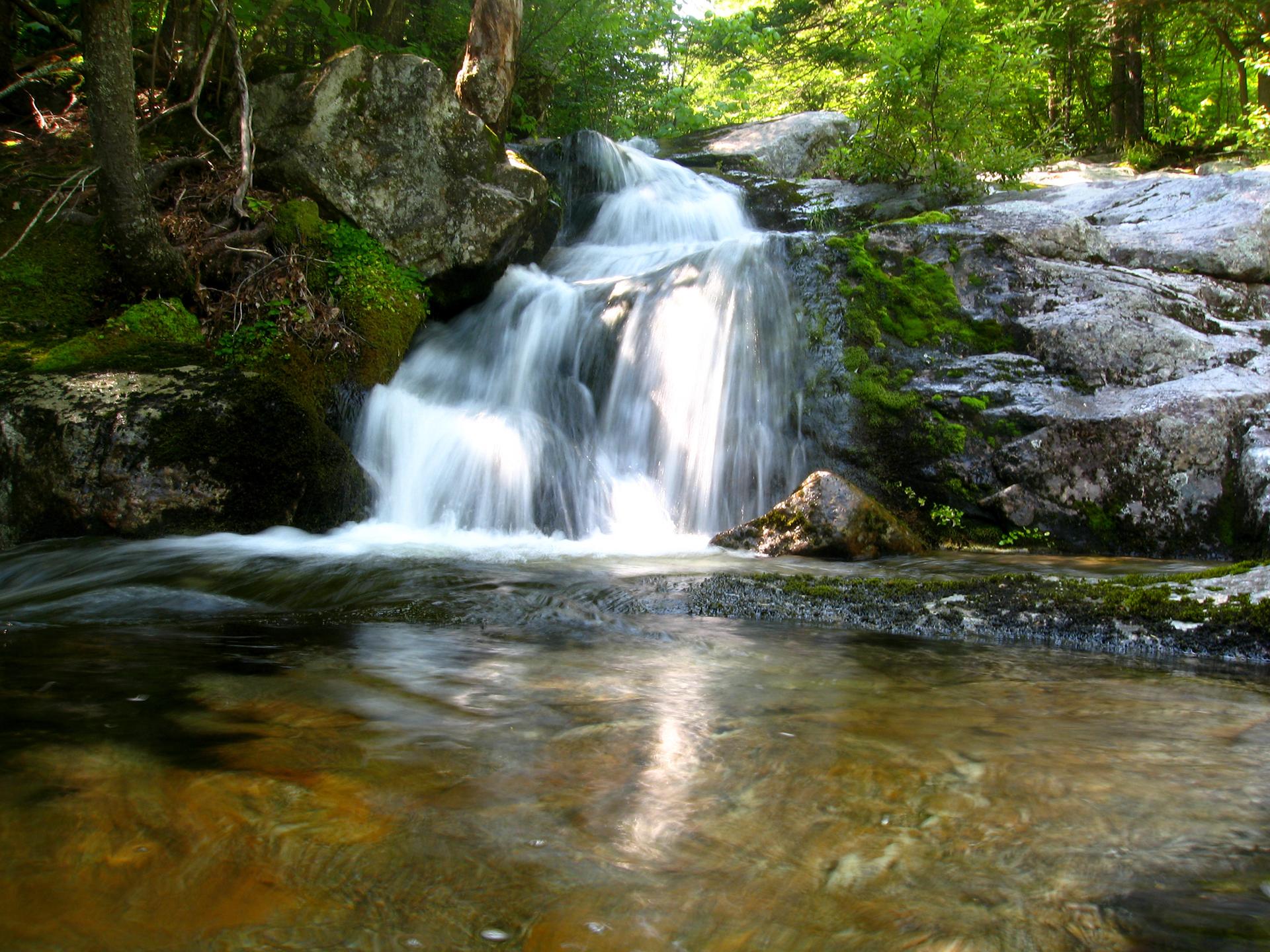A beautiful waterfall cascades over mossy rocks into a clear stream under bright light.