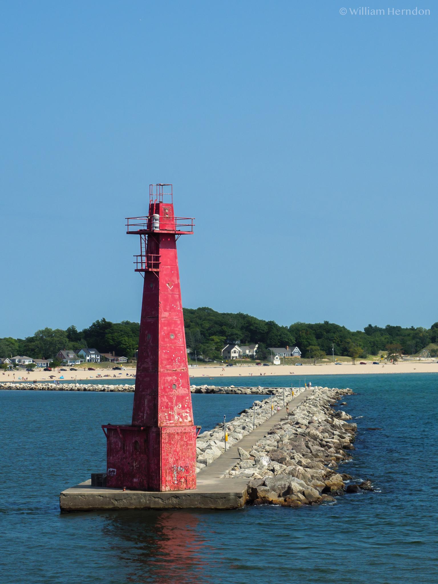 A bright red lighthouse stands on a rocky pier in blue water.