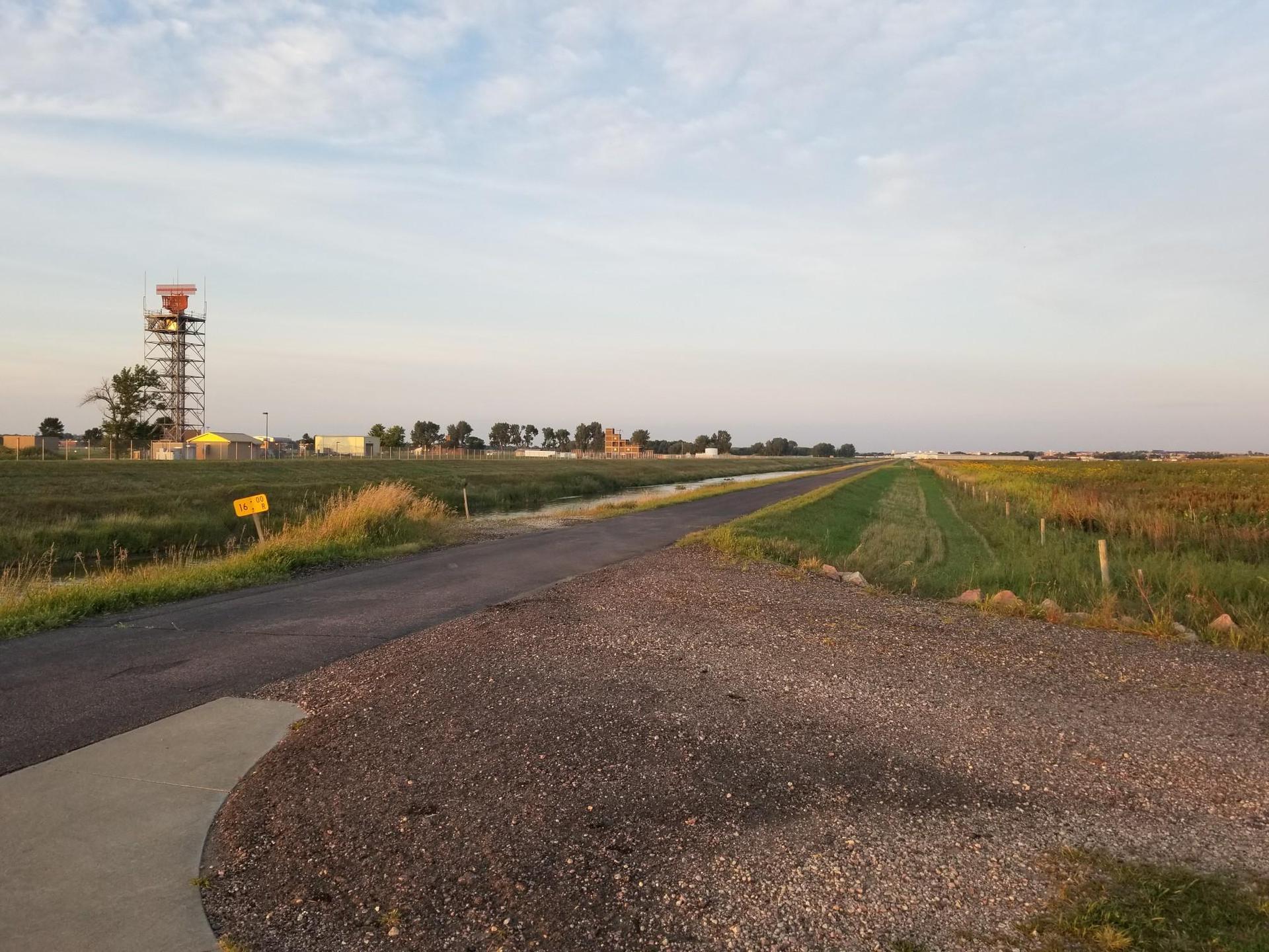 A long road through flat fields with a tower under a partly cloudy sky.