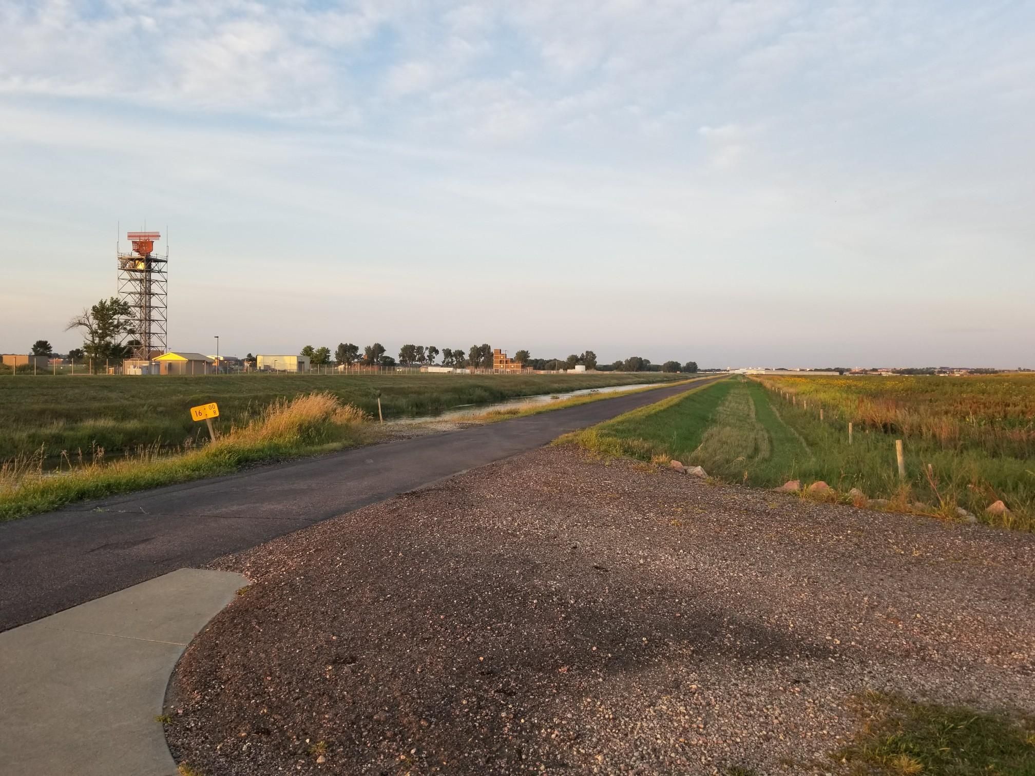 A long road through flat fields with a tower under a partly cloudy sky. - The Big Sioux River Recreation Trail and Greenway