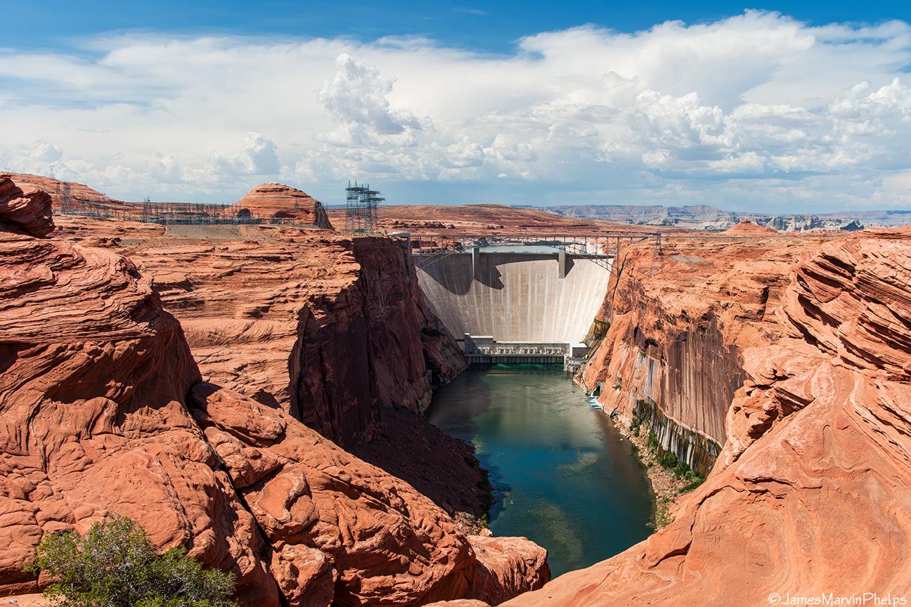 Massive concrete dam spans a canyon, river flows below, cloudy sky.
