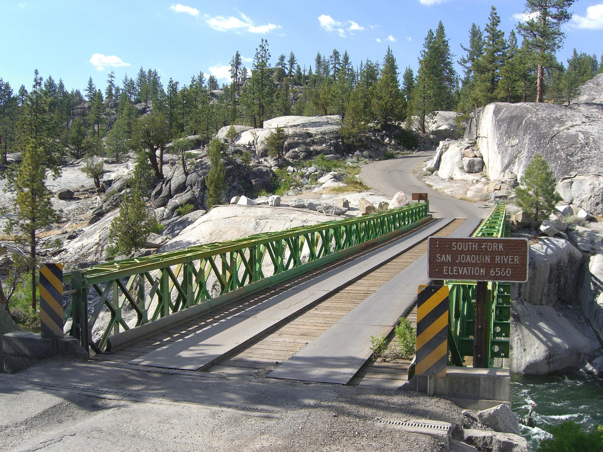 Green metal bridge over a river, surrounded by pine trees and rocky landscape.