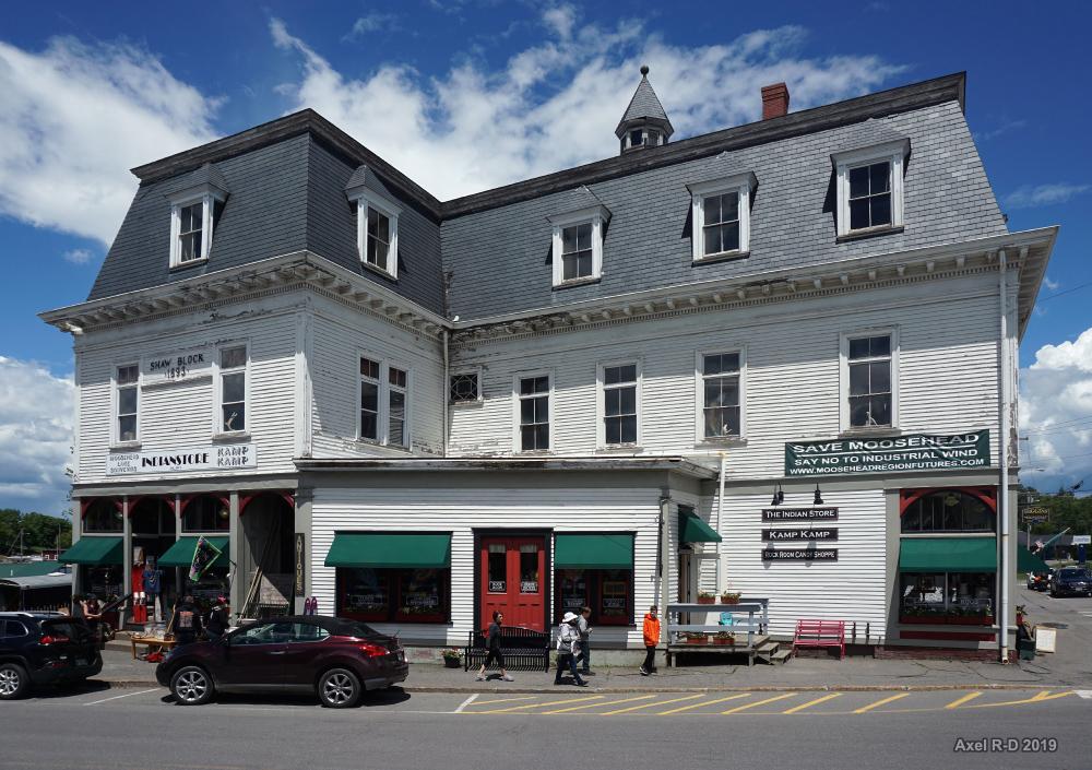 Large white building with shops under a bright, cloudy sky, people walking.