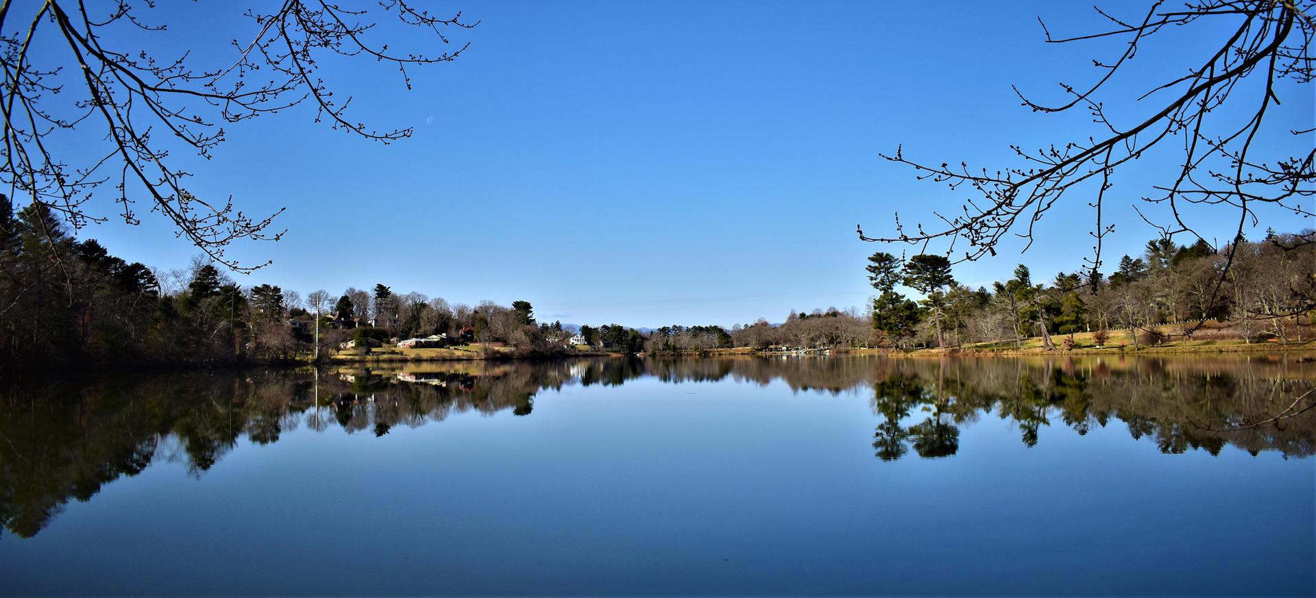 Serene lake reflecting trees and clear blue sky, framed by bare branches.