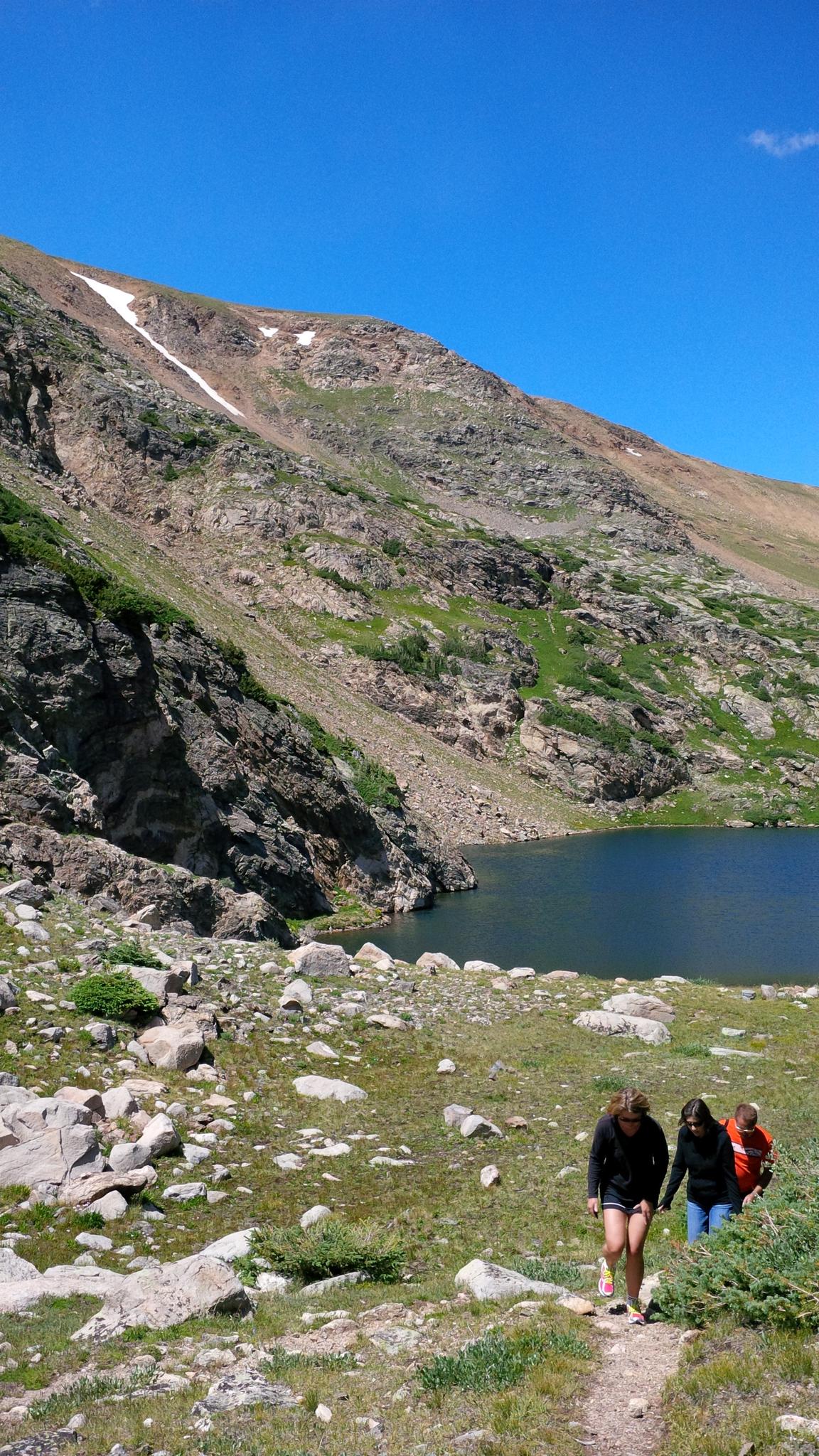 Hikers walk a path near a mountain lake under a bright blue sky.