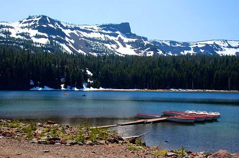 Small boats docked on a clear lake with forested shores and snow-capped mountains.