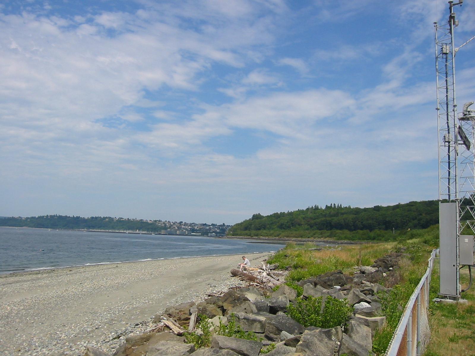 Rocky beach with calm water, distant land, and a tall metal tower.