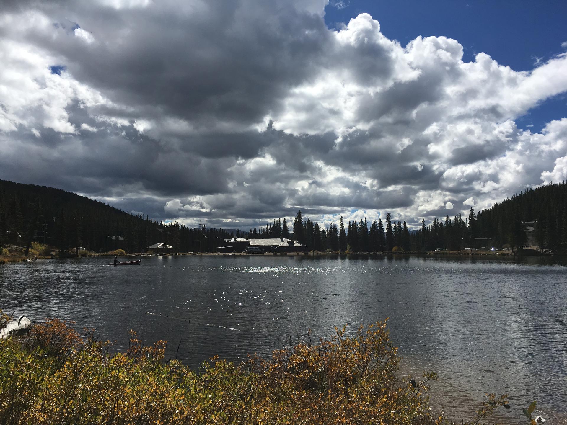 A dark lake under a dramatic cloudy sky, surrounded by forests and distant buildings.