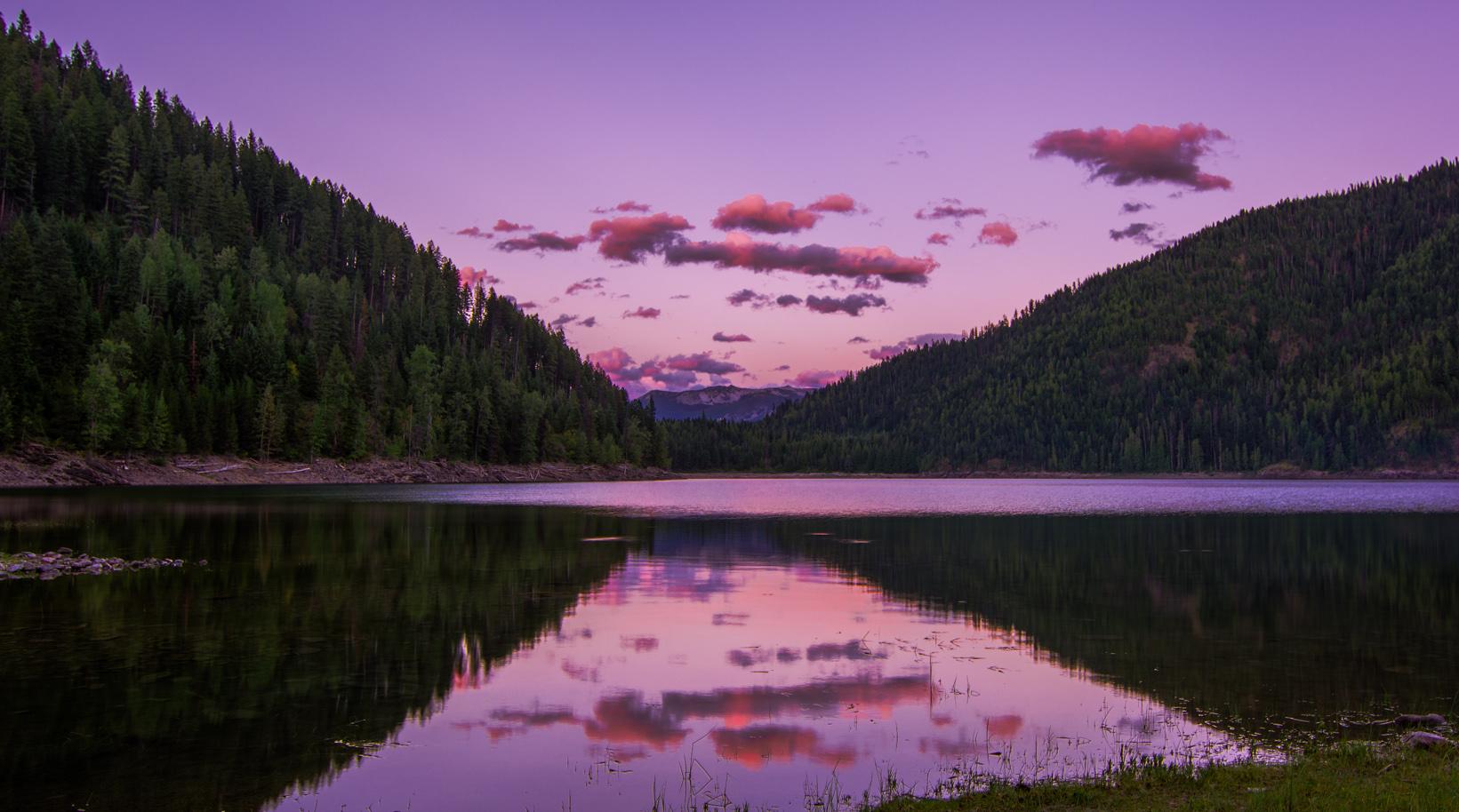 Purple sunset over a calm lake reflecting forested hills and pink clouds.