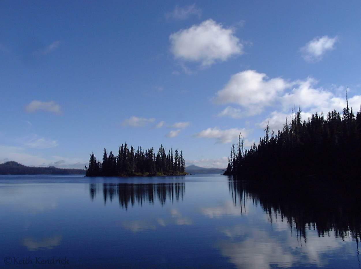 Calm lake reflecting blue sky and clouds, with dark forested islands and shore.