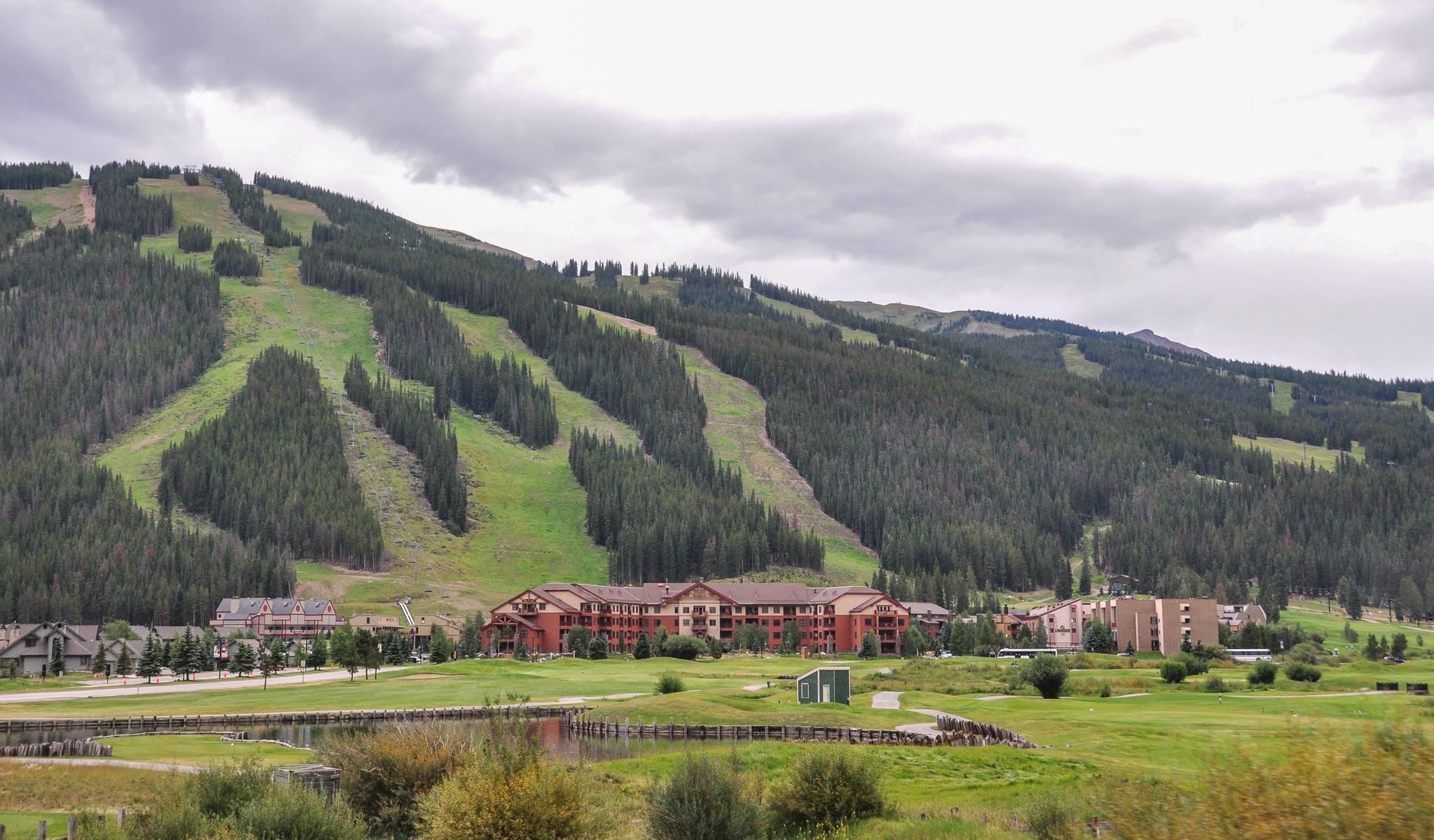 Green ski slopes above resort buildings and a pond under cloudy skies.