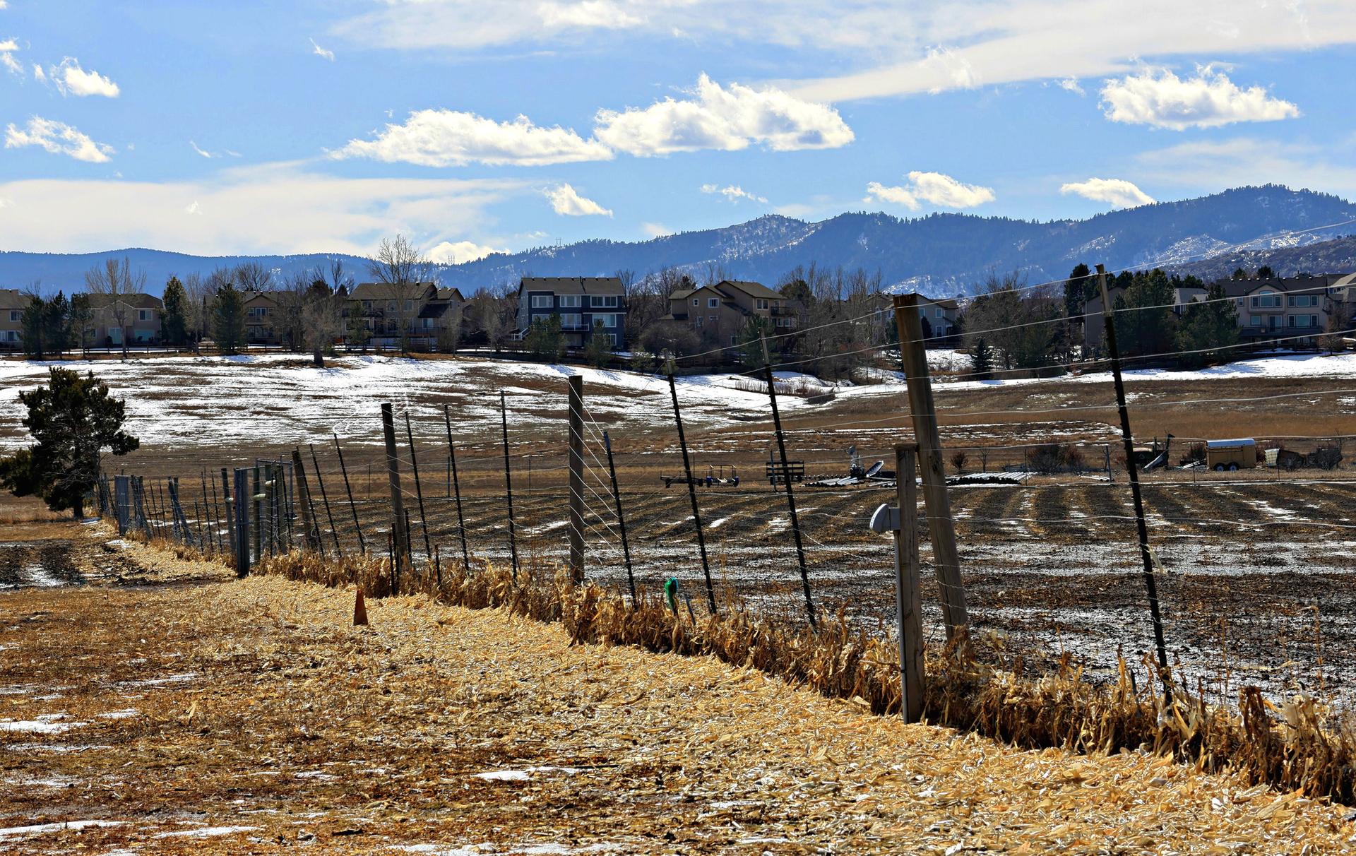 A rural field with a fence, snow patches, and distant mountains under a blue sky.