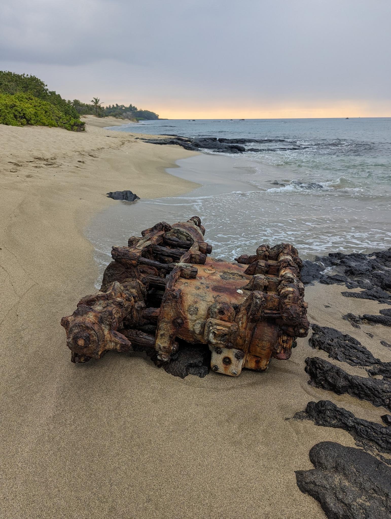 Rusty metal wreckage on a sandy beach with dark rocks and ocean waves.