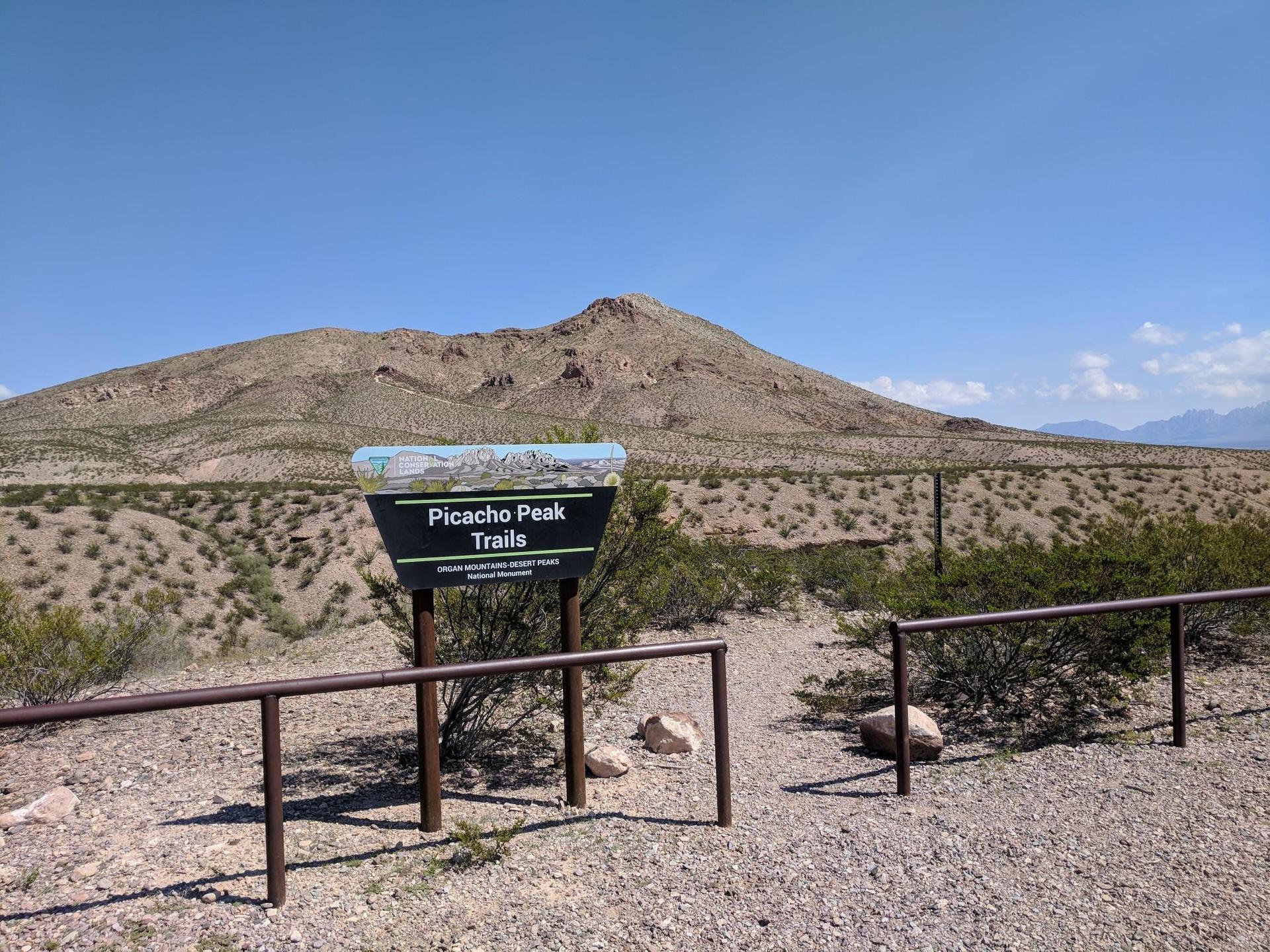 A trail sign stands before a large, arid mountain under a clear blue sky.