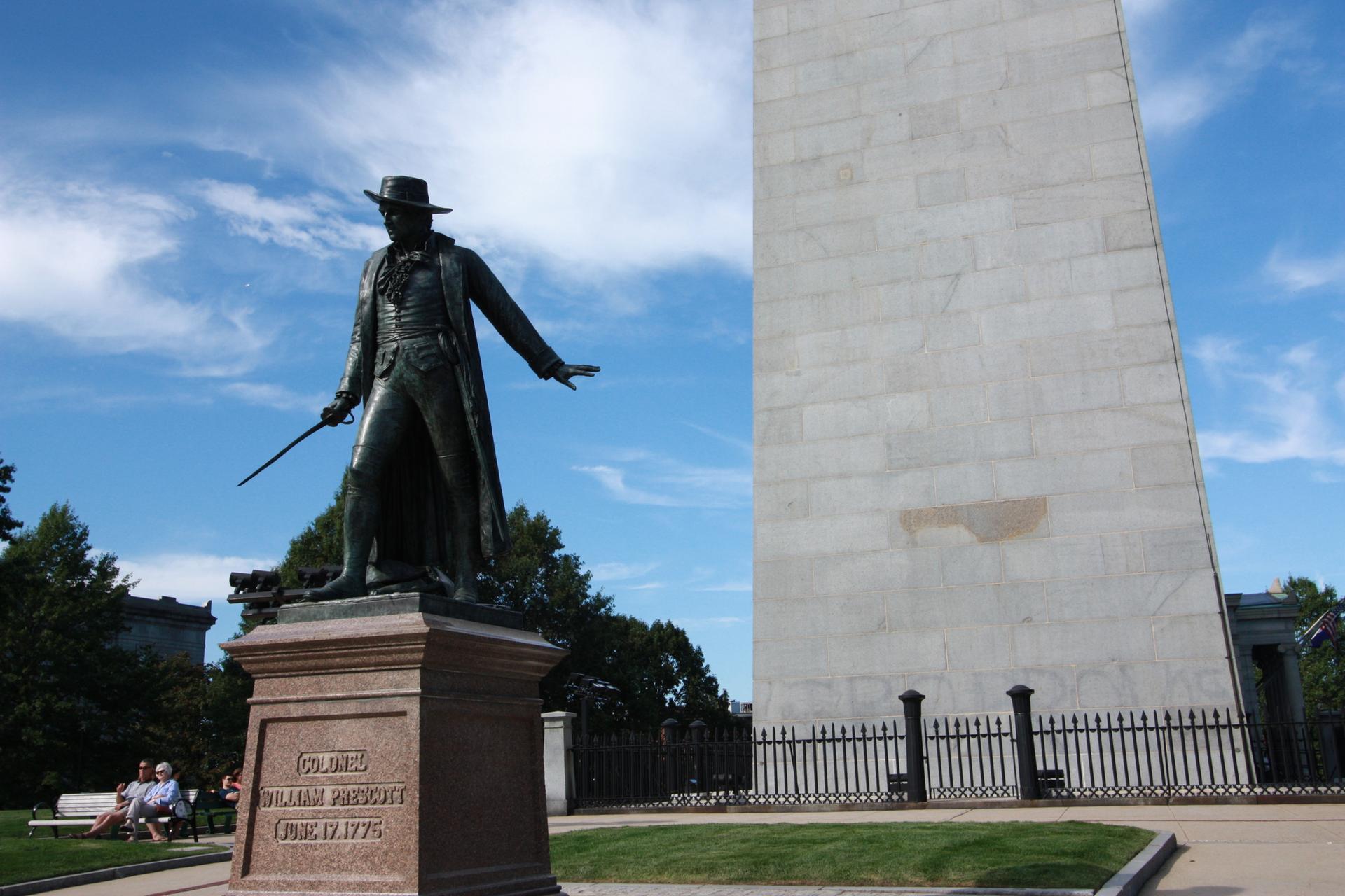 Bronze statue of a man next to a large stone obelisk on a sunny day.
