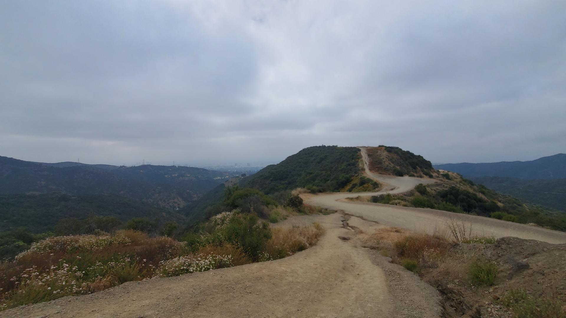 Winding dirt road on a hill with distant green valleys under an overcast sky.