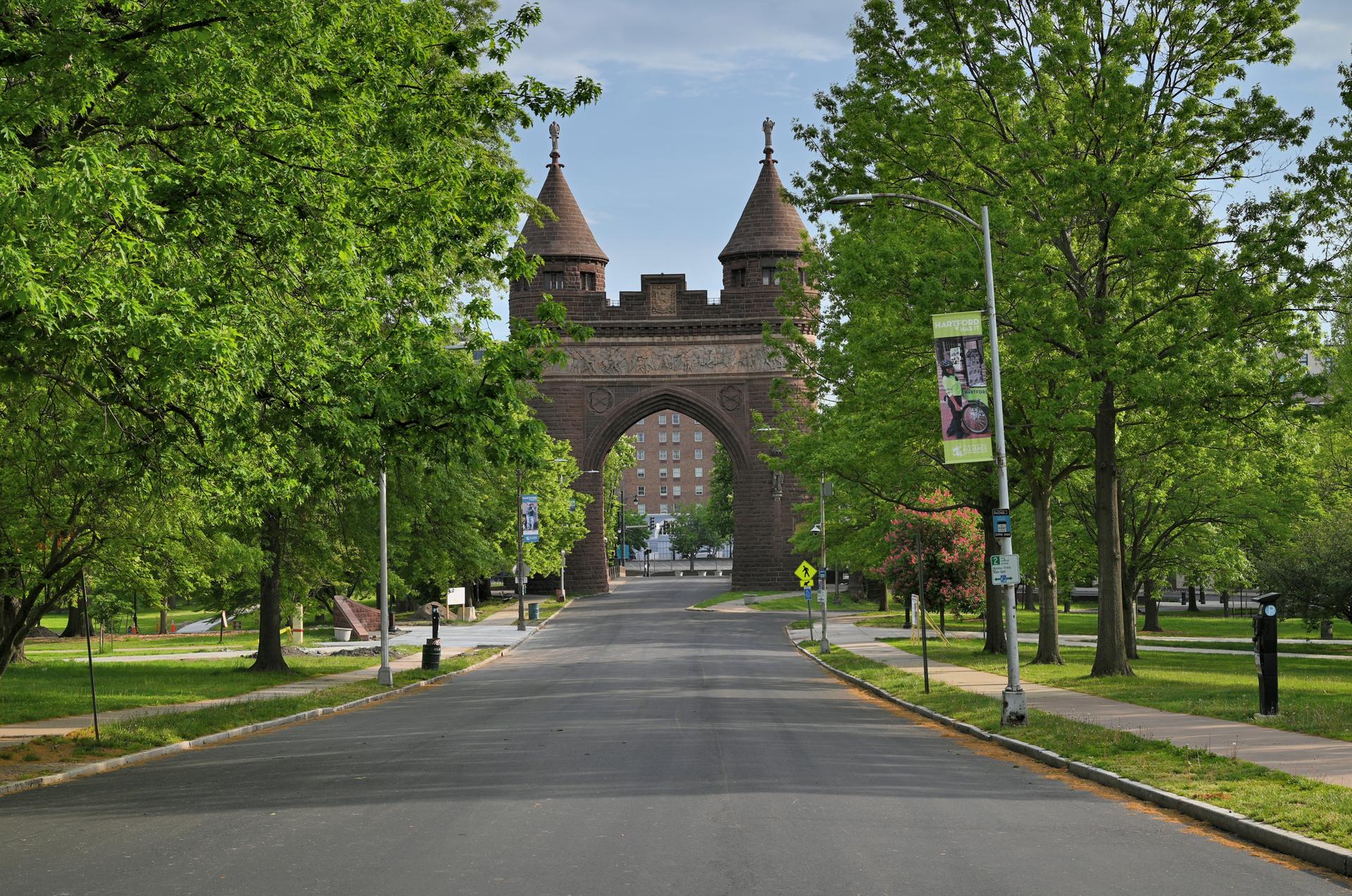 A tree-lined road leading to a large stone archway with towers on a bright day.