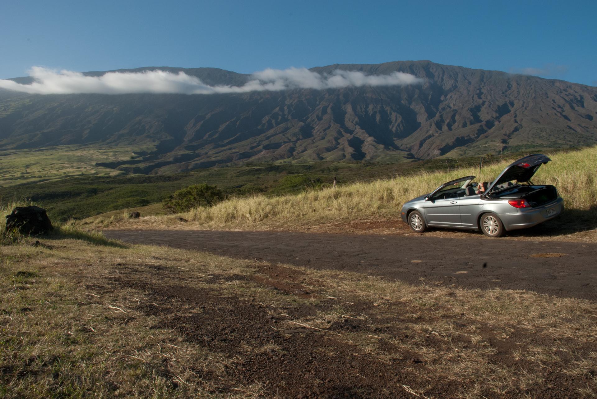 Silver convertible car parked on a dirt road, with large mountains and clouds.