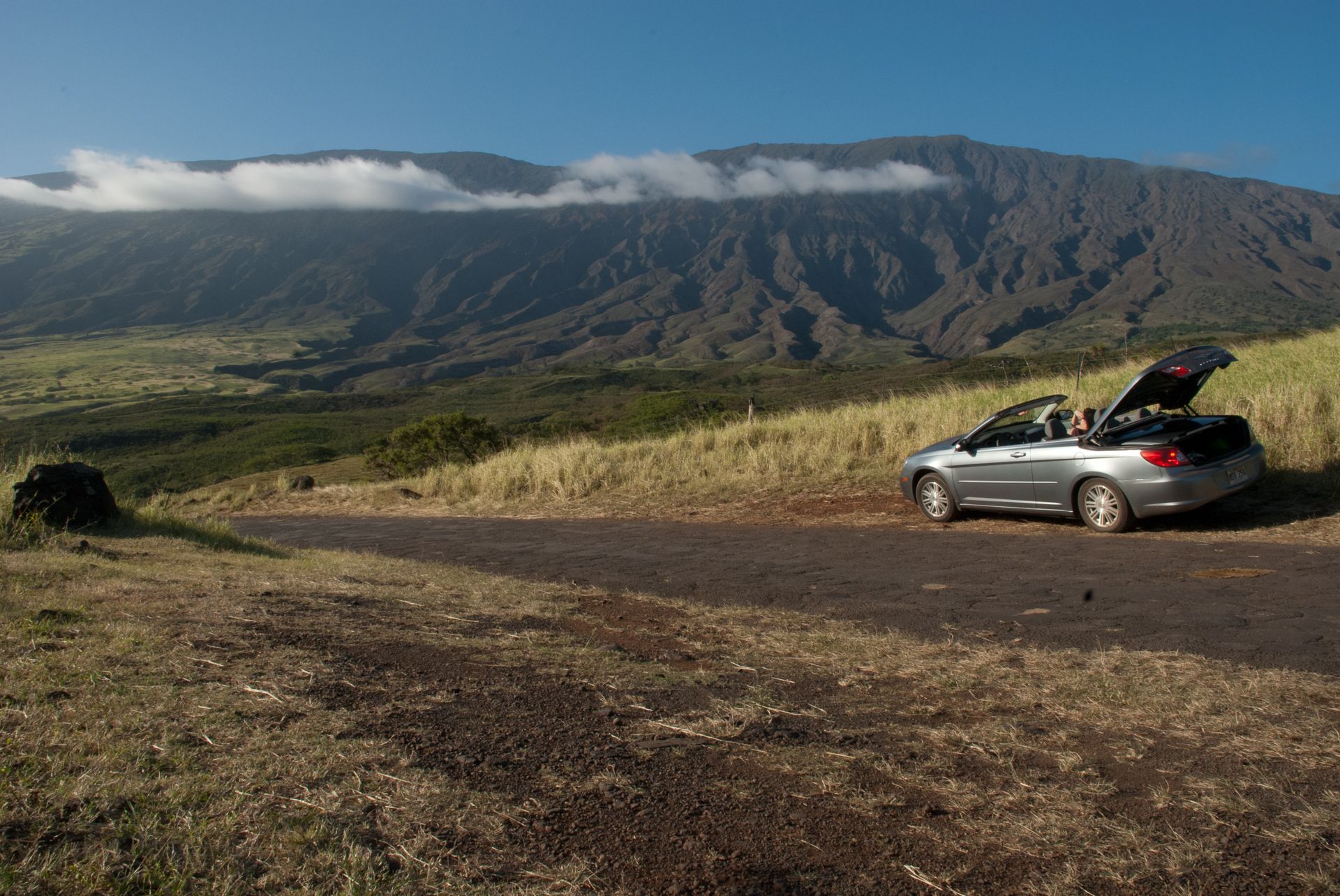 Silver convertible car parked on a dirt road, with large mountains and clouds. - Pi'ilani Highway