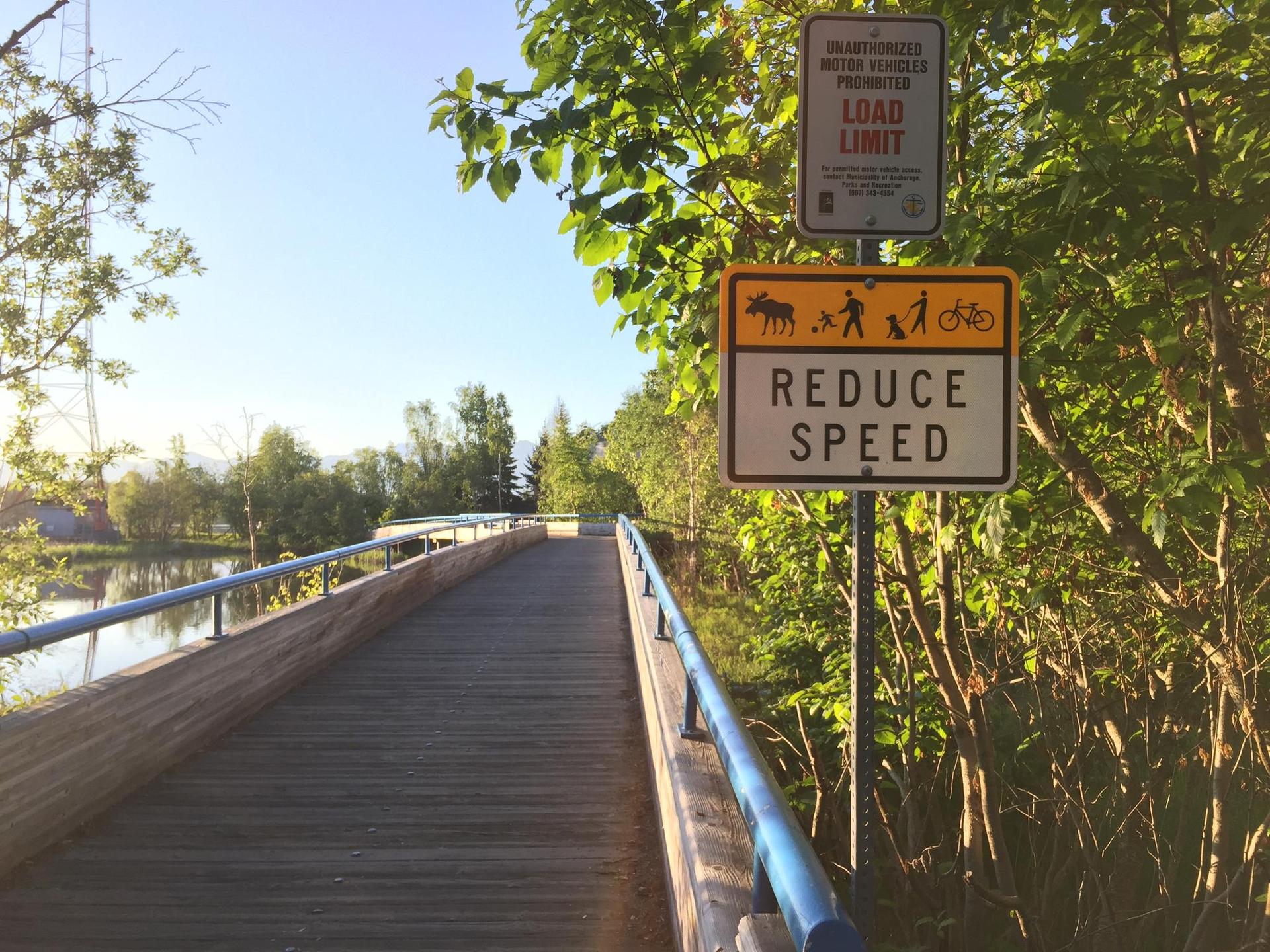 Wooden boardwalk next to a lake with a "Reduce Speed" sign, sunny.