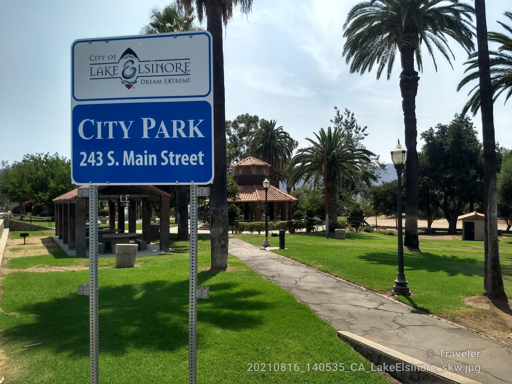 Blue park sign in a sunny park with palm trees and a paved path.