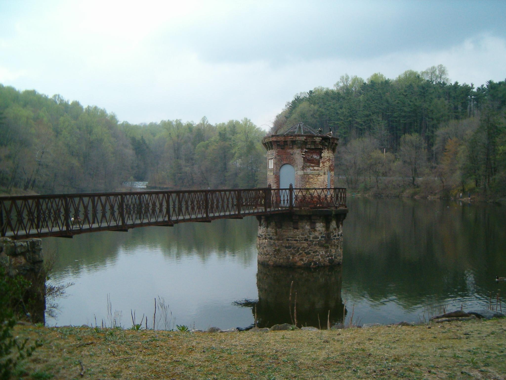 Old stone tower and metal bridge over a calm lake, surrounded by green trees.
