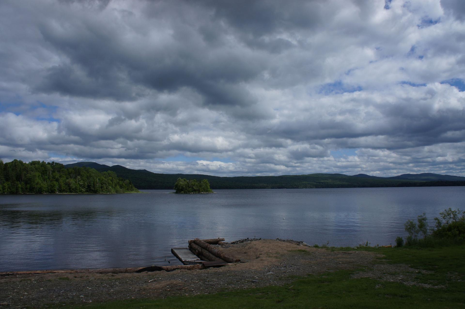 Cloudy sky over a lake with an island, forested hills, and a small dock.