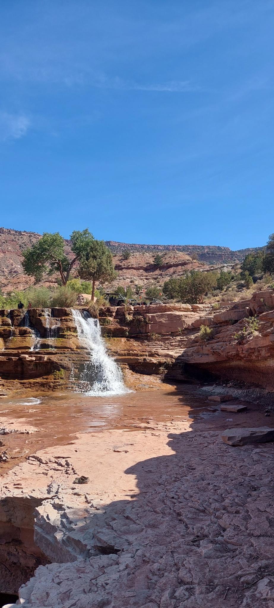 A waterfall cascades into a red rock pool under a bright blue sky.
