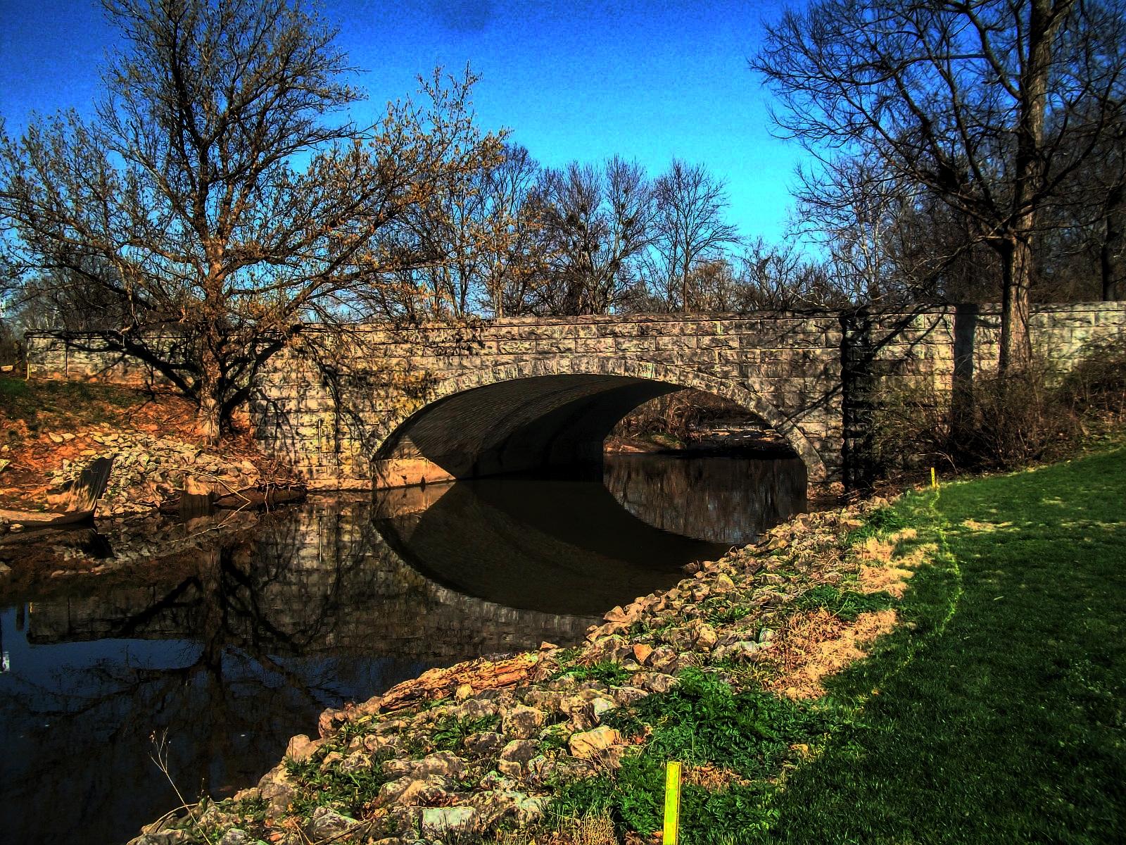 A stone arch bridge spans dark water, reflecting bare trees under a blue sky.