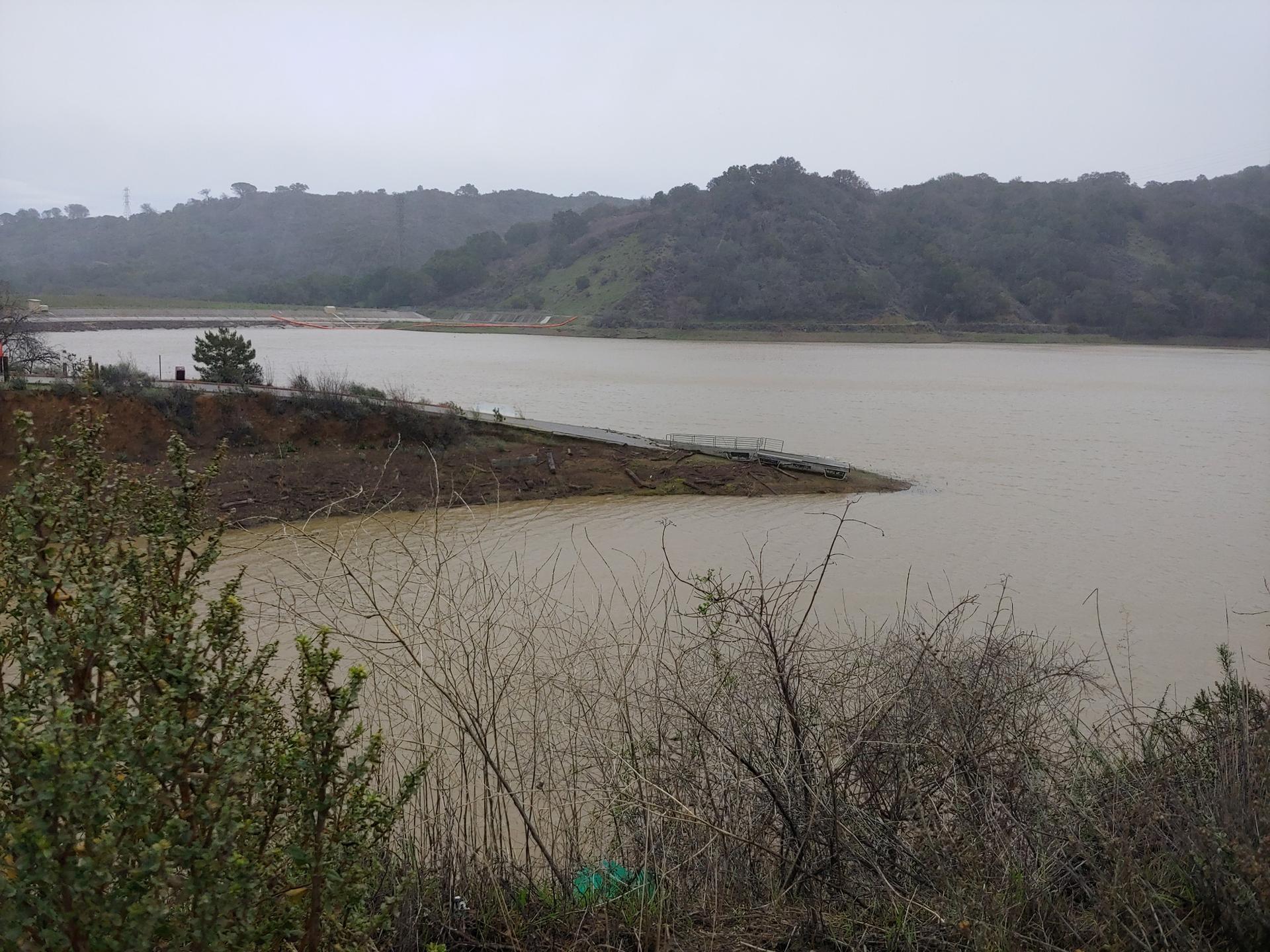 Overcast view of a murky lake with hills and dry bushes in the foreground.