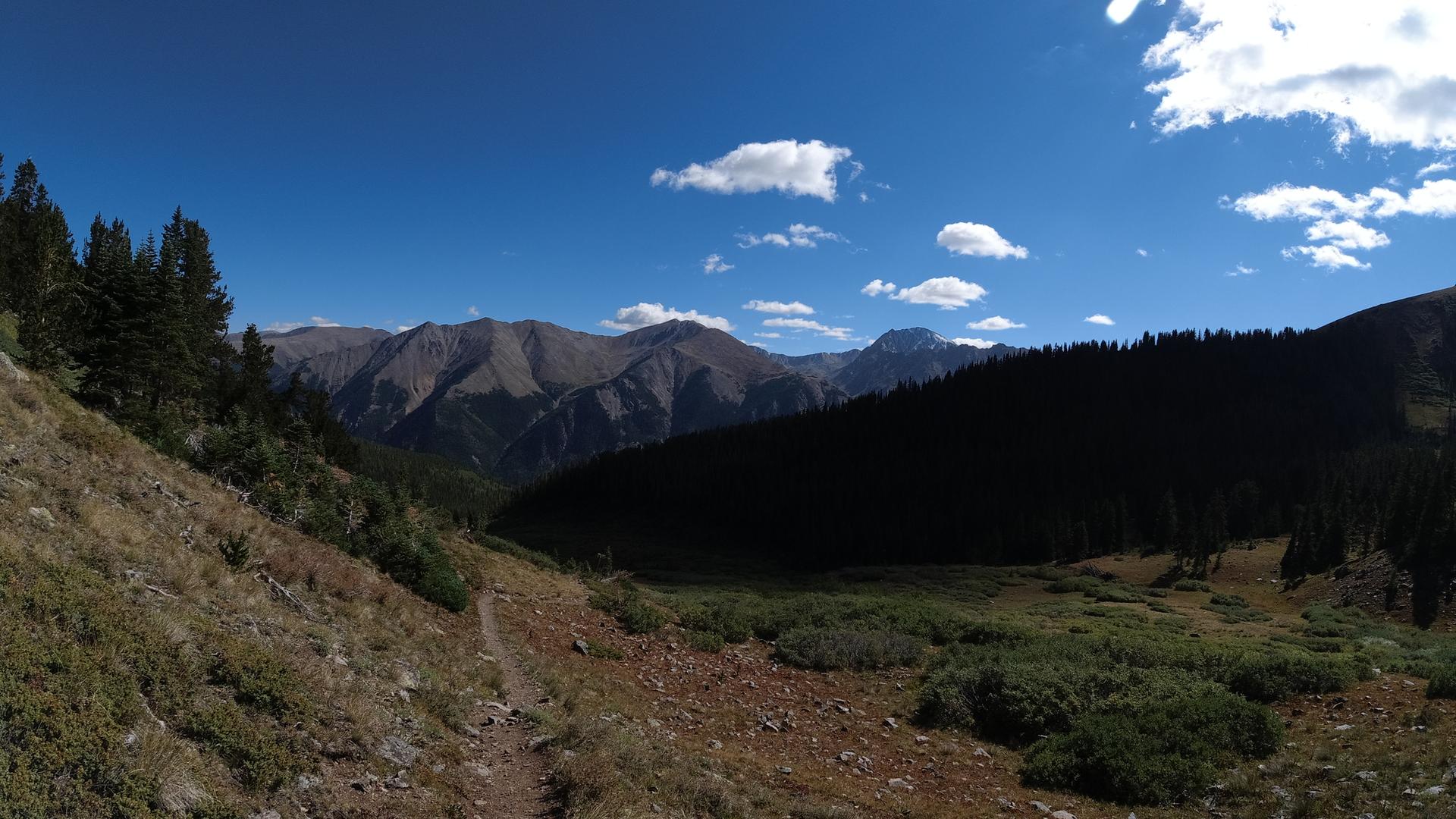 A dirt path winds through a mountain valley with forested slopes and distant peaks.