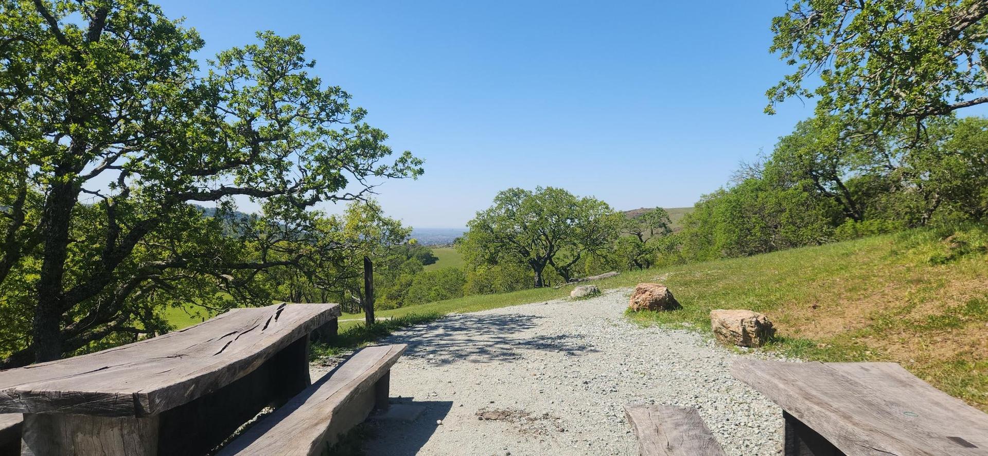 Wooden picnic tables beside a gravel path overlooking green hills on a sunny day.