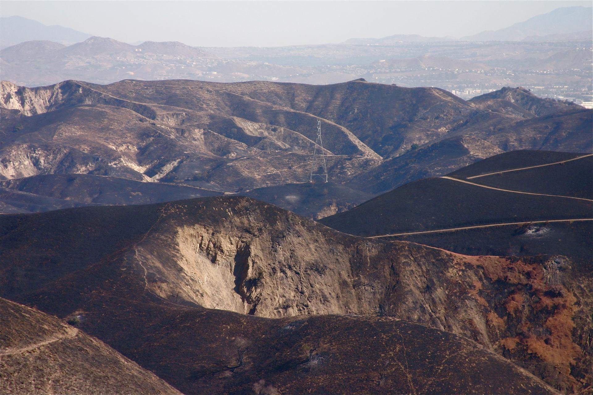 Expansive view of rugged, burnt mountains with distant city haze under bright light.