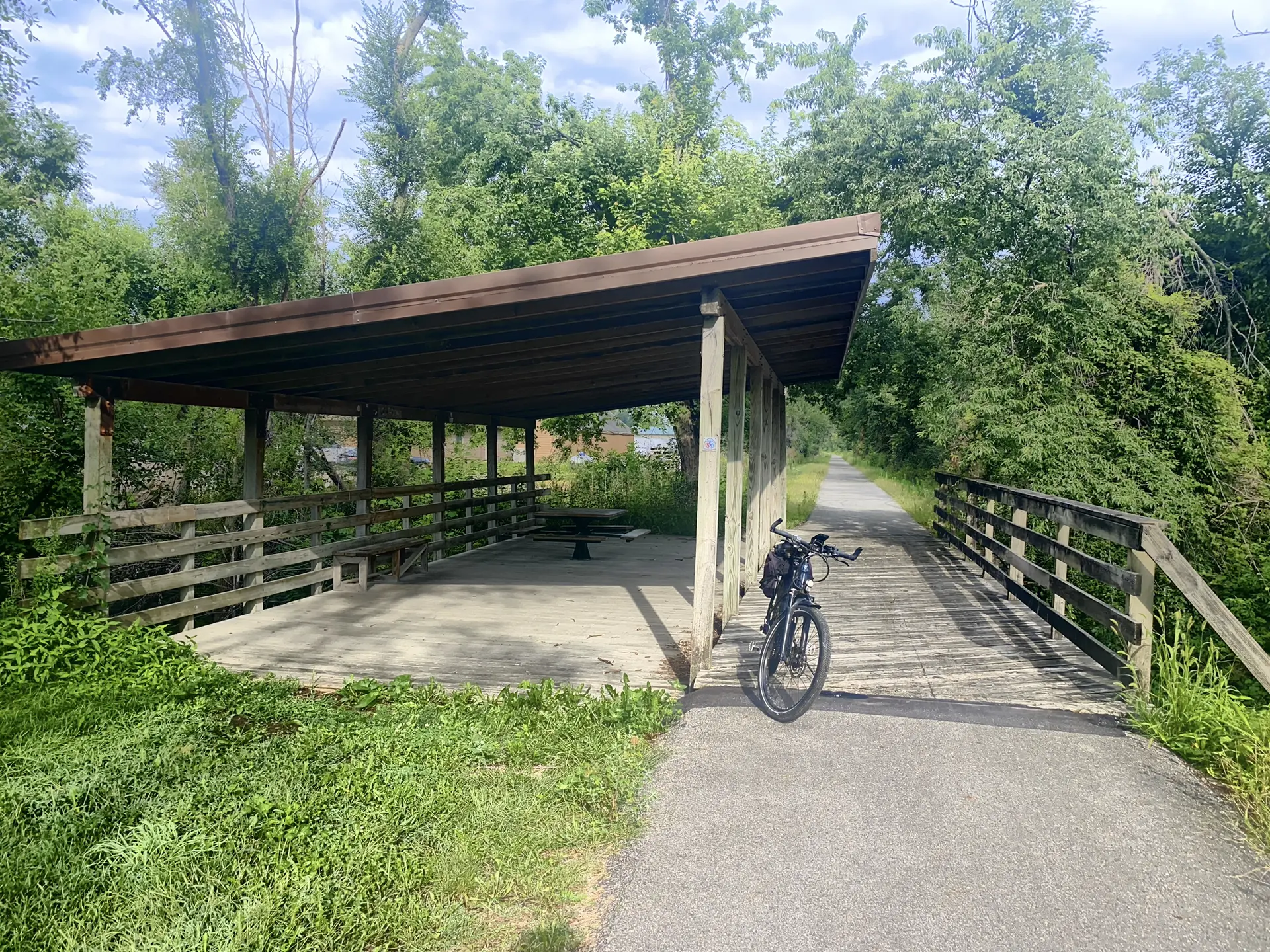A bicycle parked next to a wooden picnic shelter on a paved path.