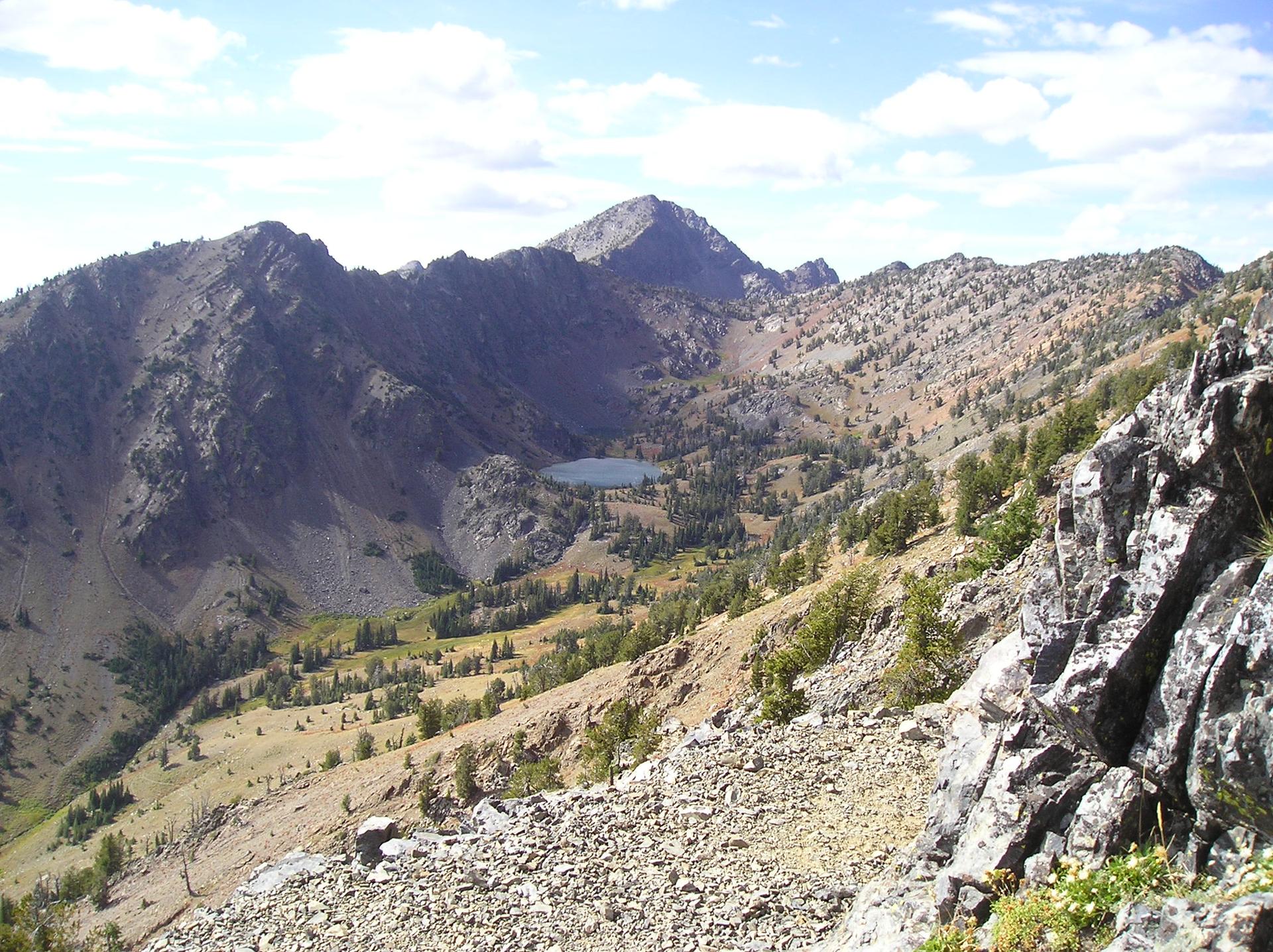 Rugged mountains, a small lake, and a valley under a bright, cloudy sky.