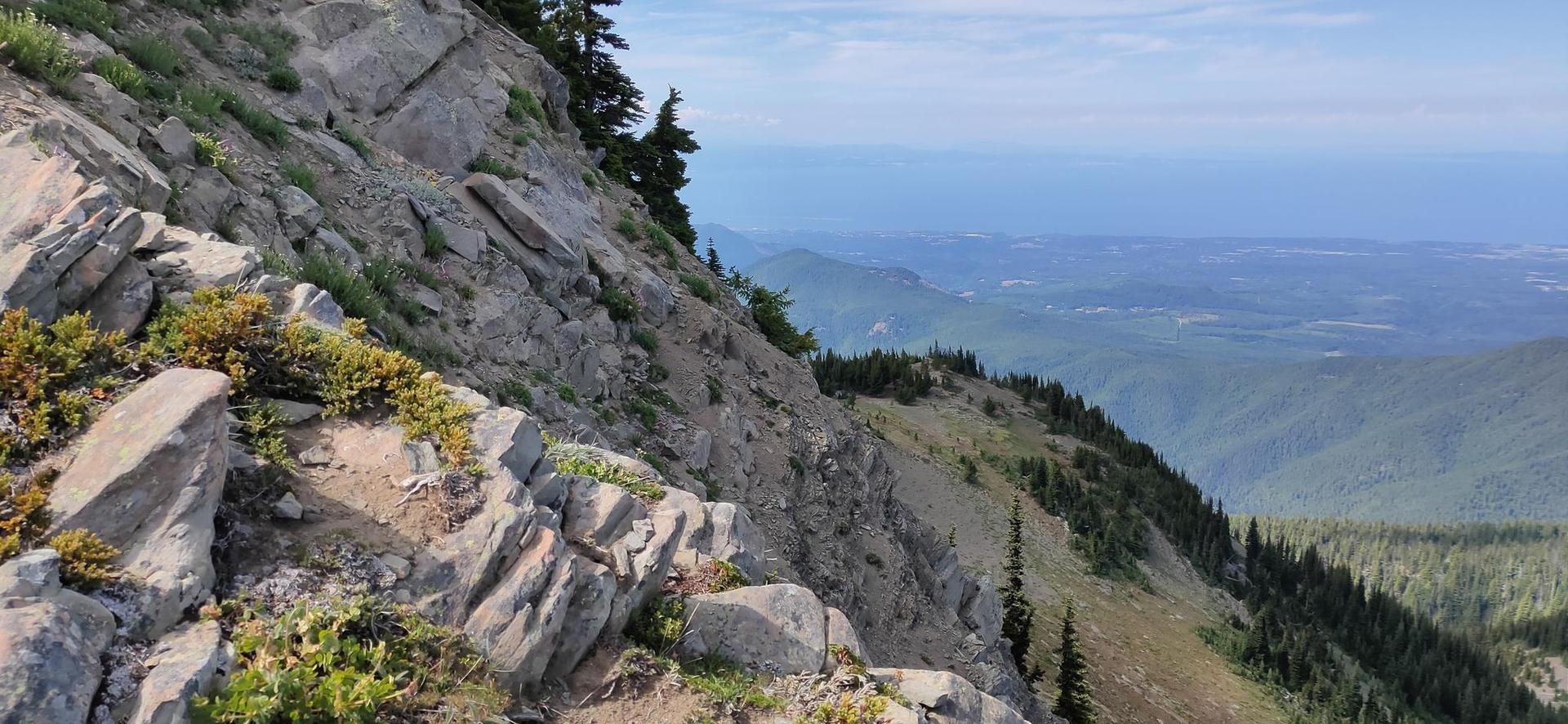 Steep rocky mountain slope overlooking a vast valley and distant water.