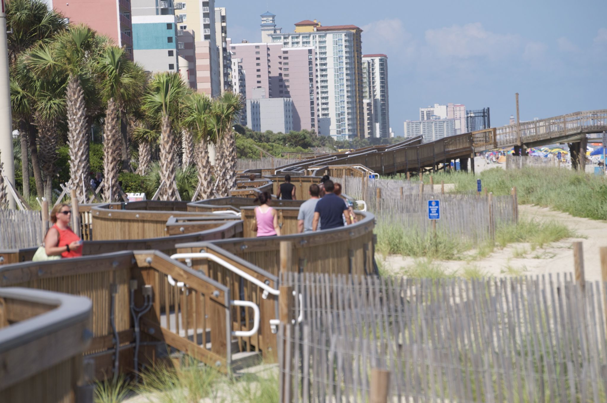 People walk on a wooden boardwalk with city buildings in the background. - Myrtle Beach loop from Myrtle Beach International Airport
