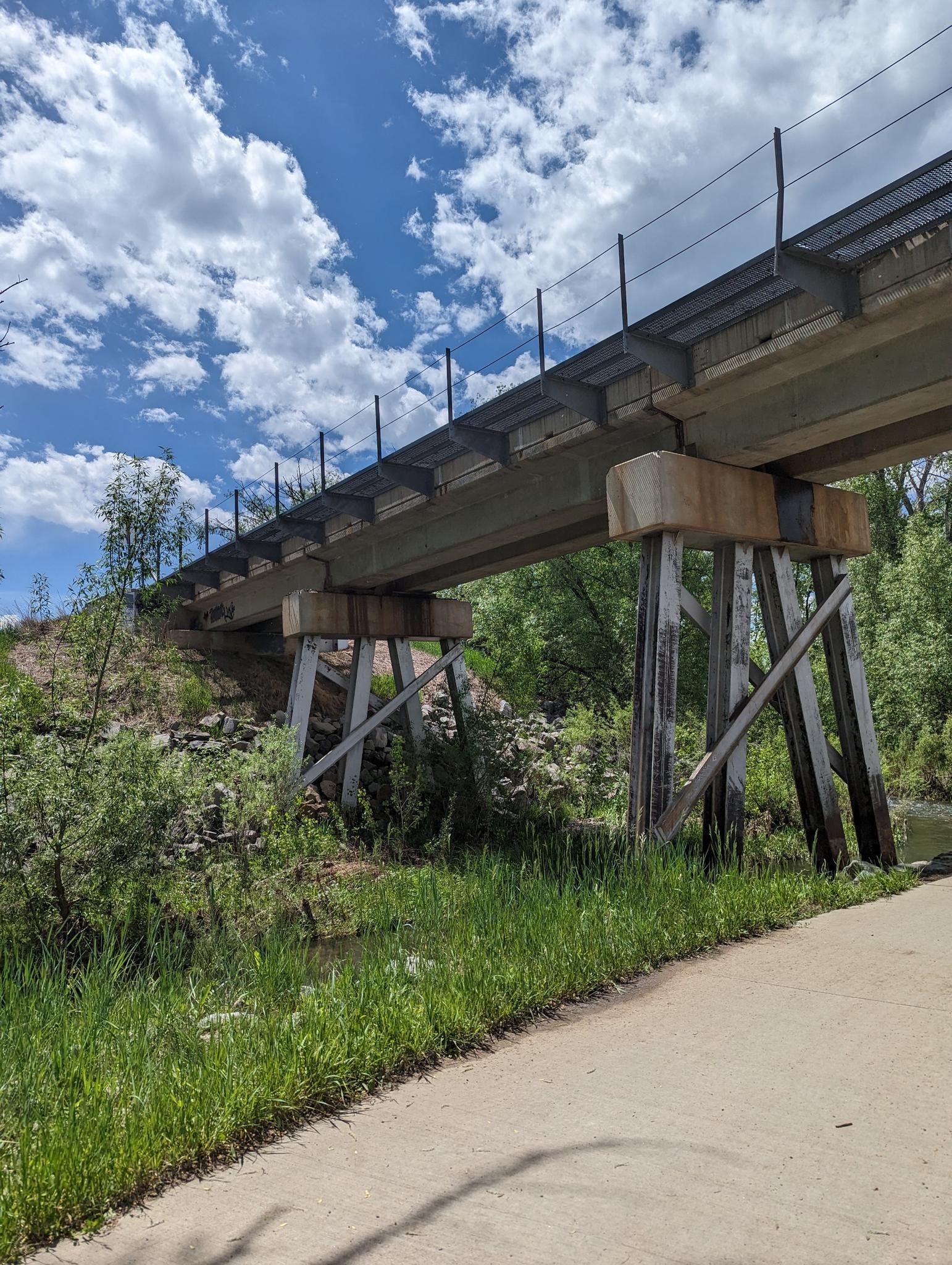 A large concrete bridge spans over water and green foliage under a cloudy sky.